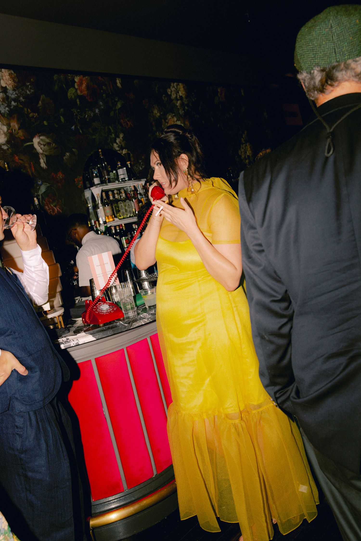 Woman in yellow gown talks on red telephone at a vibrant bar during evening party.