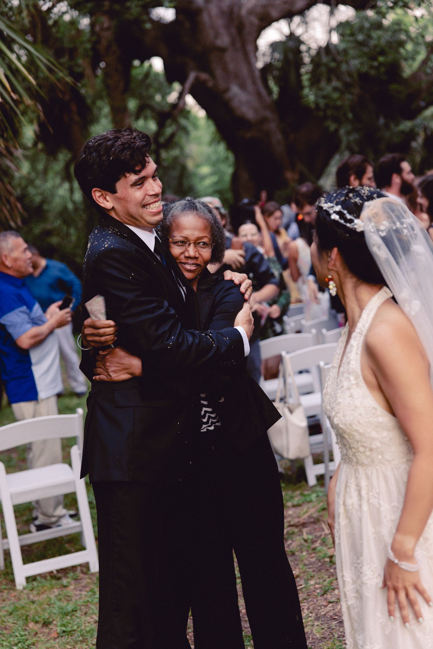 Groom embraces elderly guest while bride watches during joyful outdoor wedding ceremony recessional.