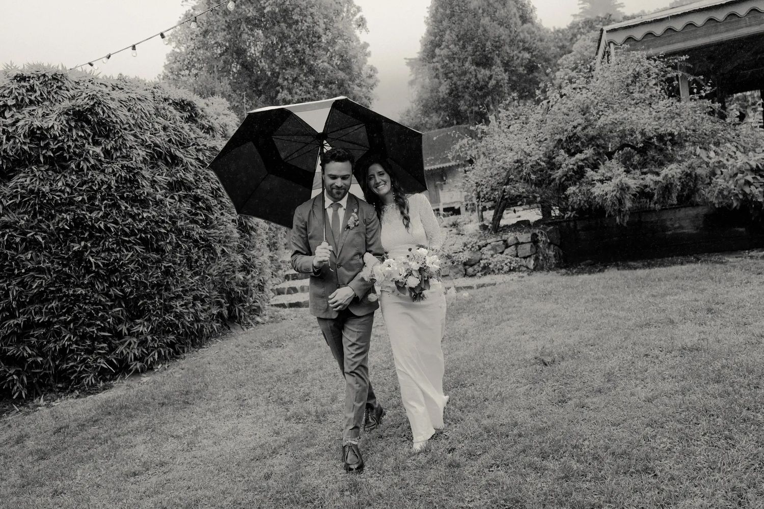 Black and white photo of newlyweds walking through garden in rain sharing an umbrella with bouquet.