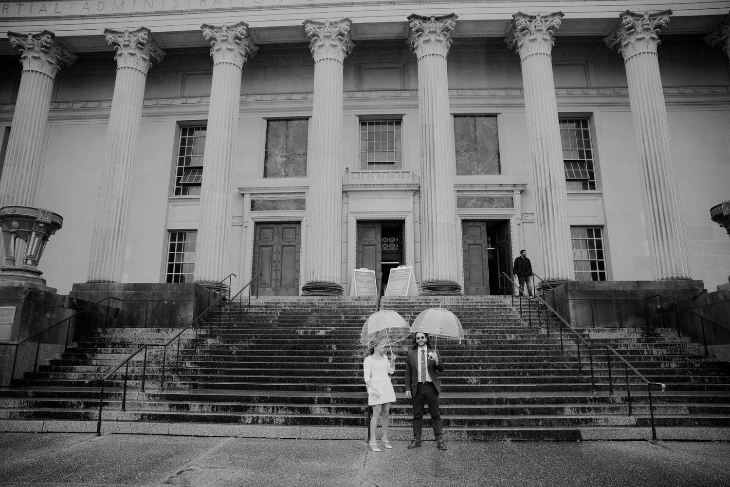 Black and white photo of couple with umbrella standing before grand columned courthouse building.