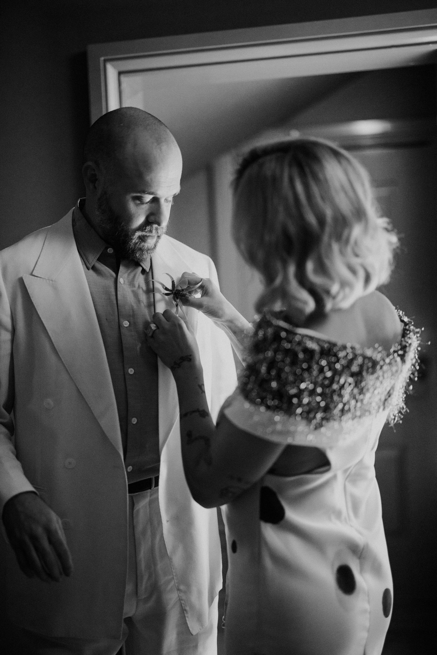 Black and white photo of bride pinning boutonniere on groom in white suit before wedding ceremony.