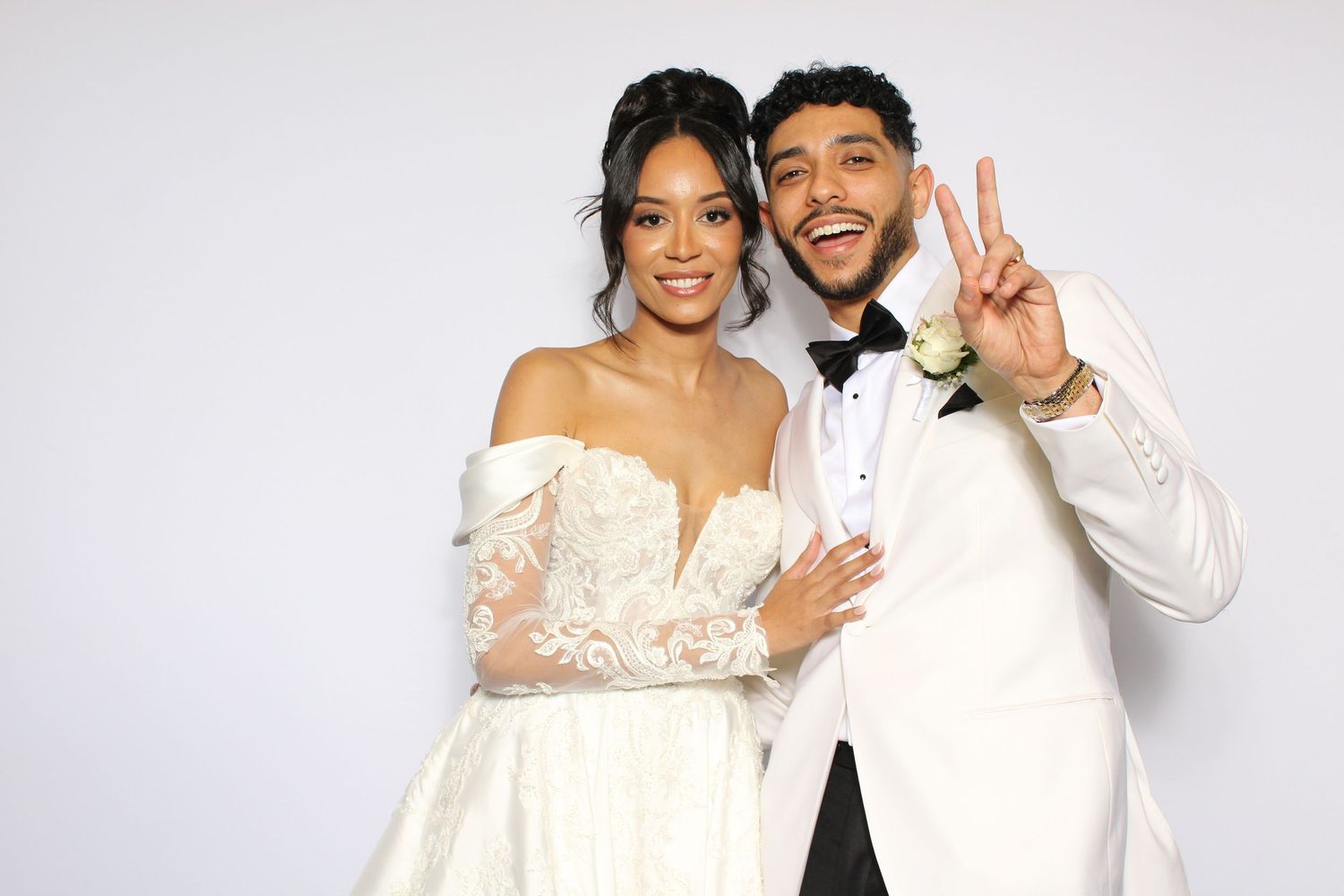 Couple in wedding attire posing for photo booth picture, bride in lace gown and groom in white tuxedo flashing peace sign.
