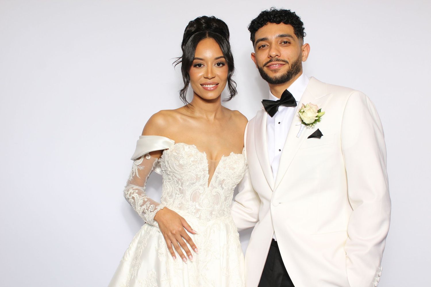 Elegant couple posing at wedding, bride in lace off-shoulder gown, groom in white tuxedo with boutonniere.