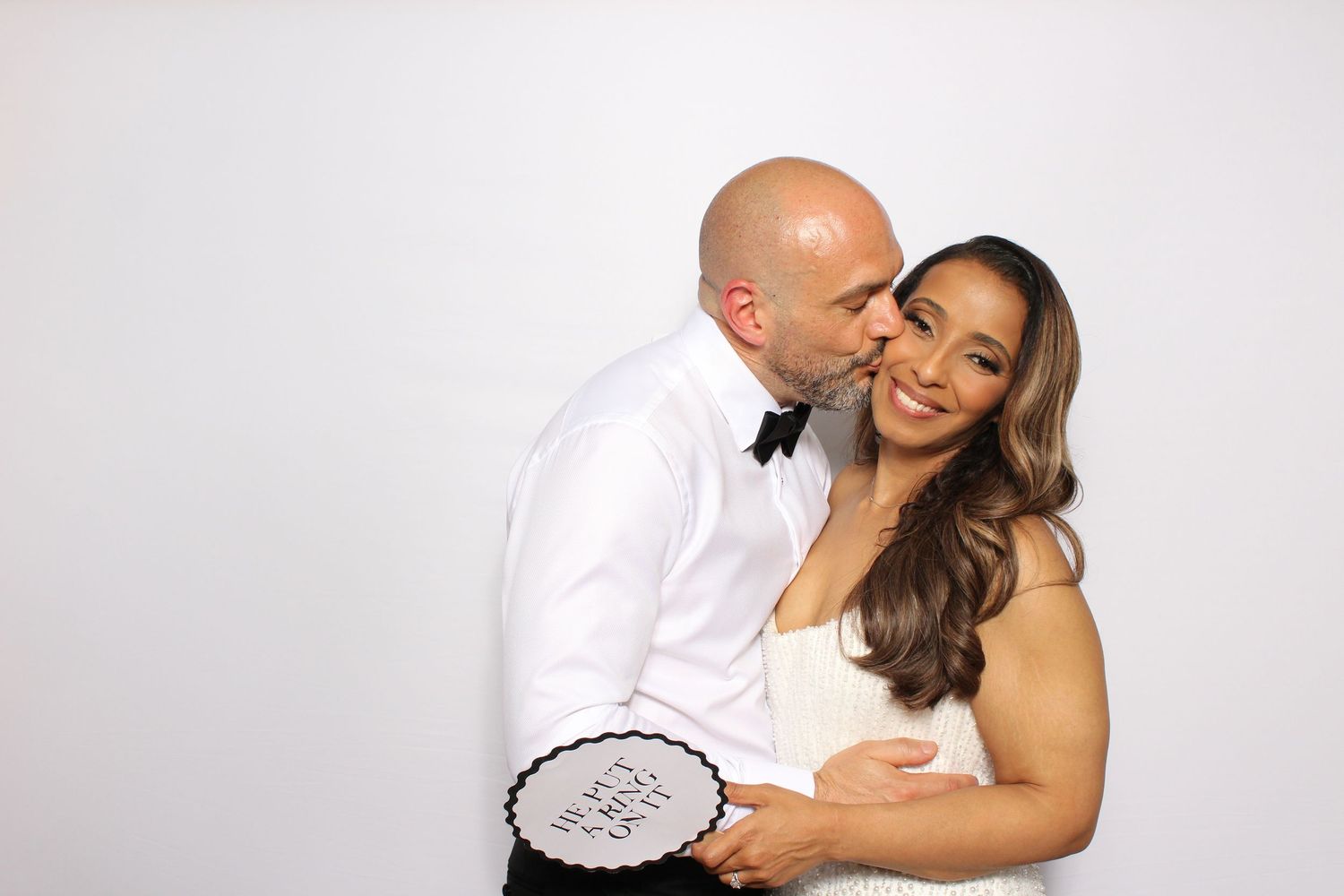 Newlywed couple posing at wedding photo booth, groom kissing bride on cheek while she smiles holding a sign.