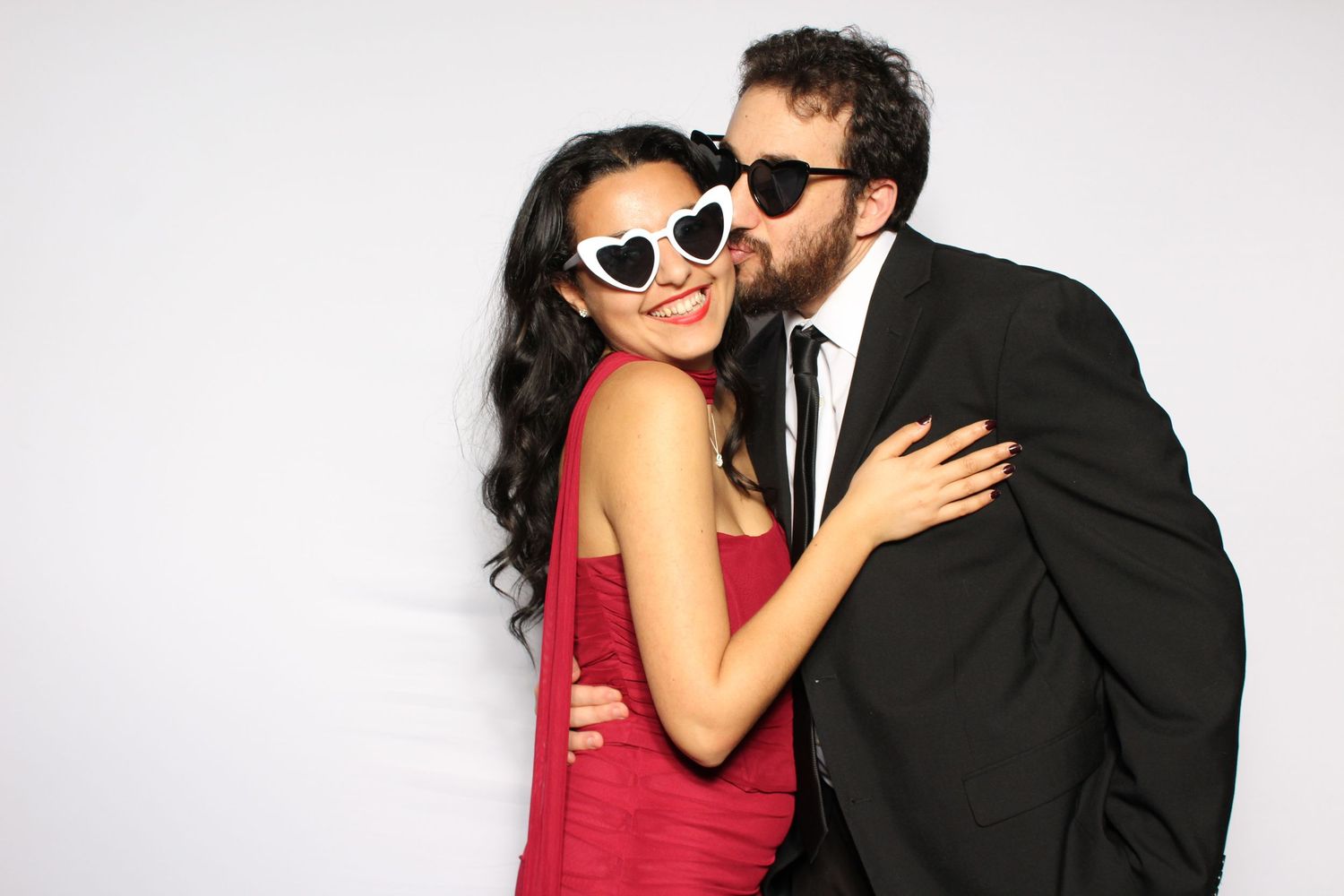 Couple posing in photo booth, woman in red dress with heart sunglasses, man in black suit kissing her cheek.