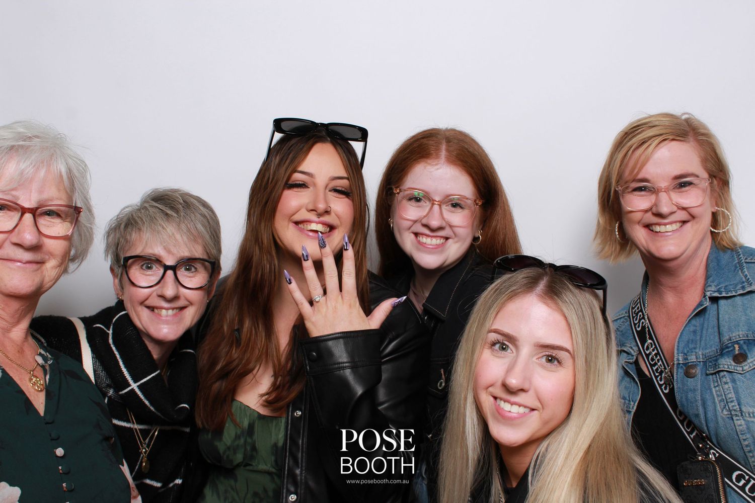 A group of people smile together in a black and white photo booth image, with one person showing off a ring.