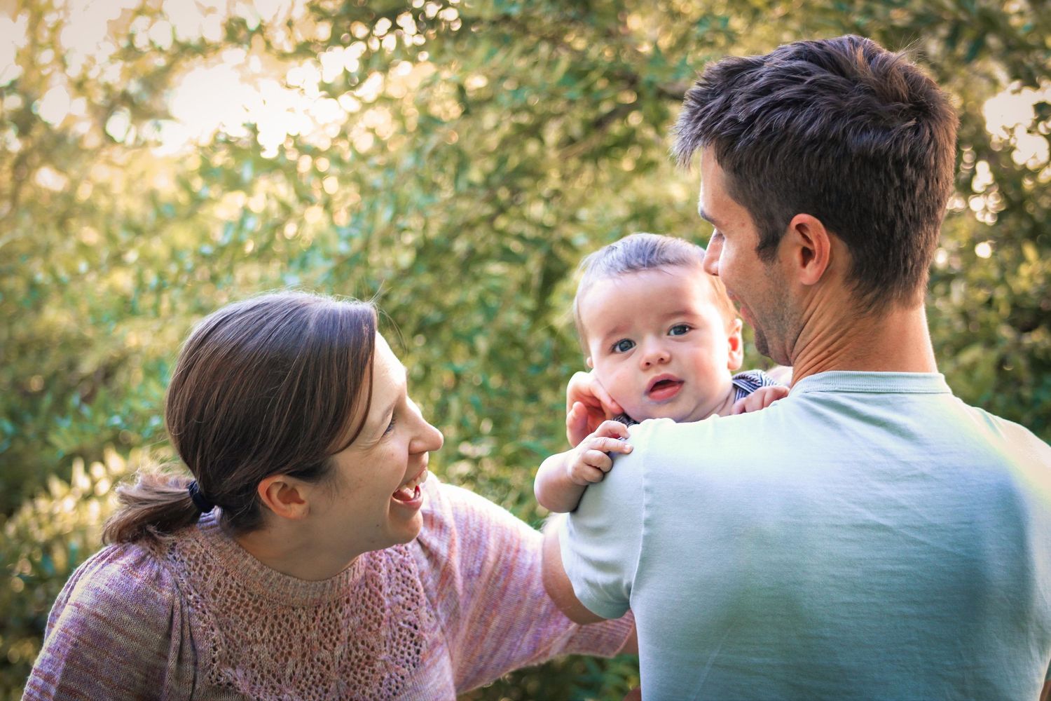 Baby smiling with mom and dad looking at the child with trees in the background