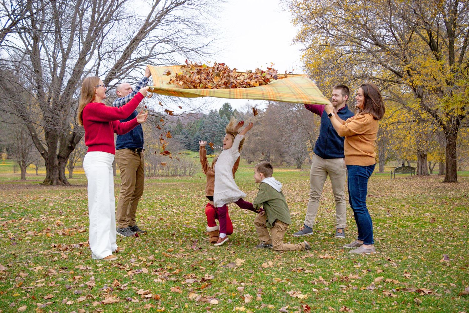 Four adults tossing a blanket with leaves on it while three kids jump underneath it