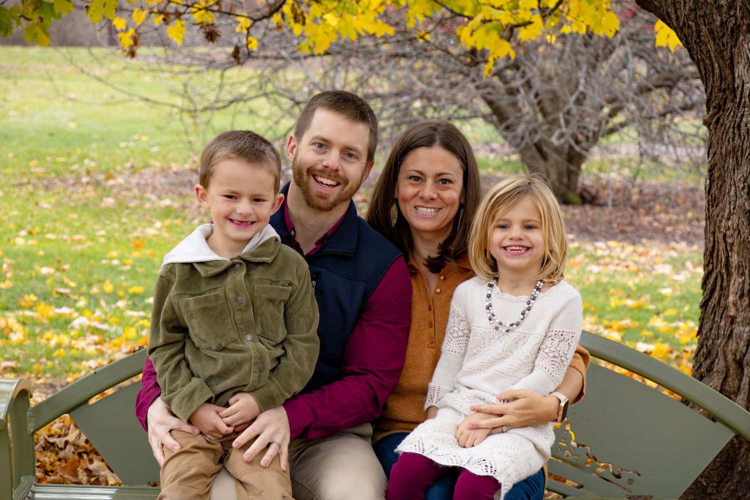 A family of four smiling at the camera while sitting on a green bench under a tree