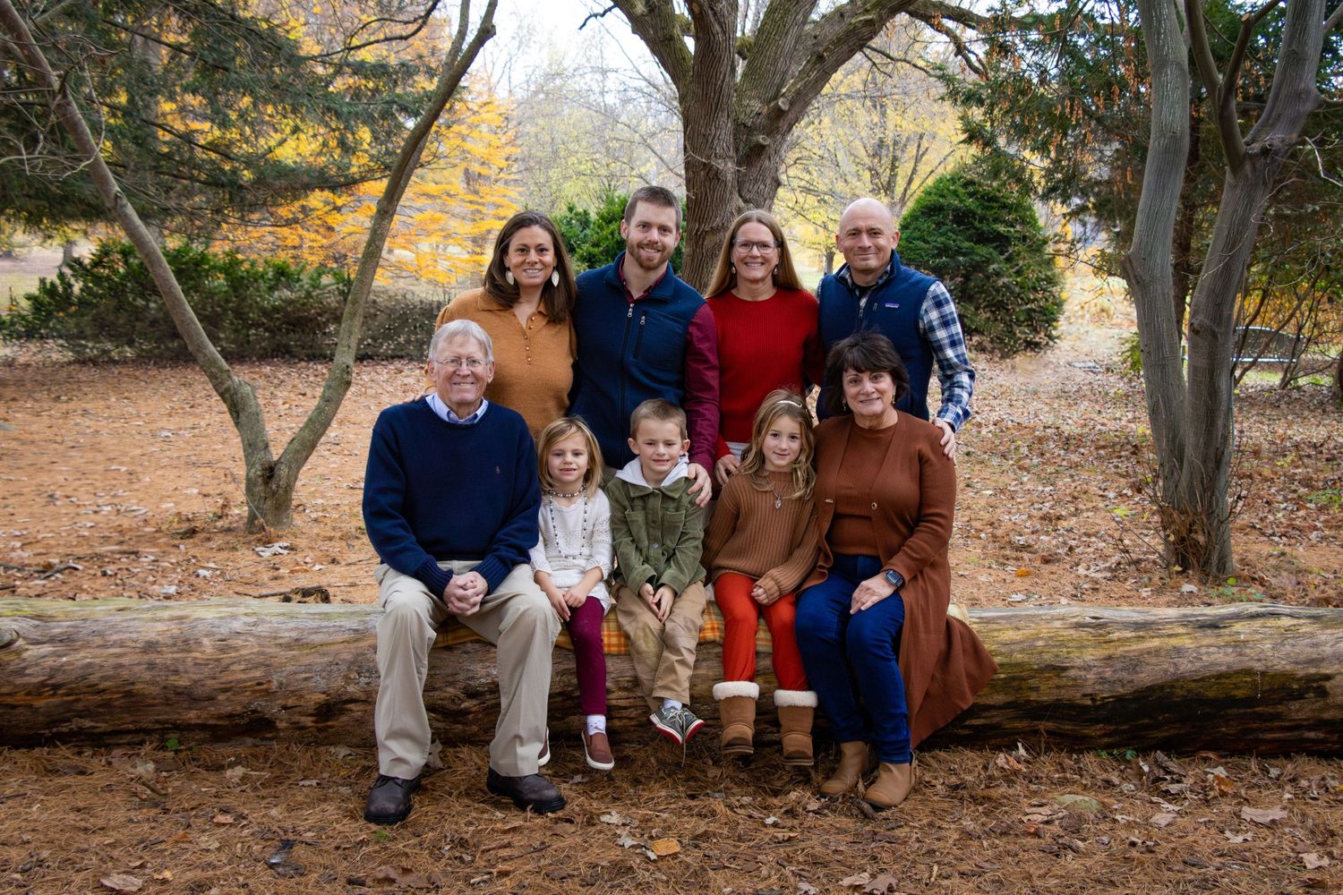 Family of nine smiles in an open field with some sitting on a long wooden log as a bench