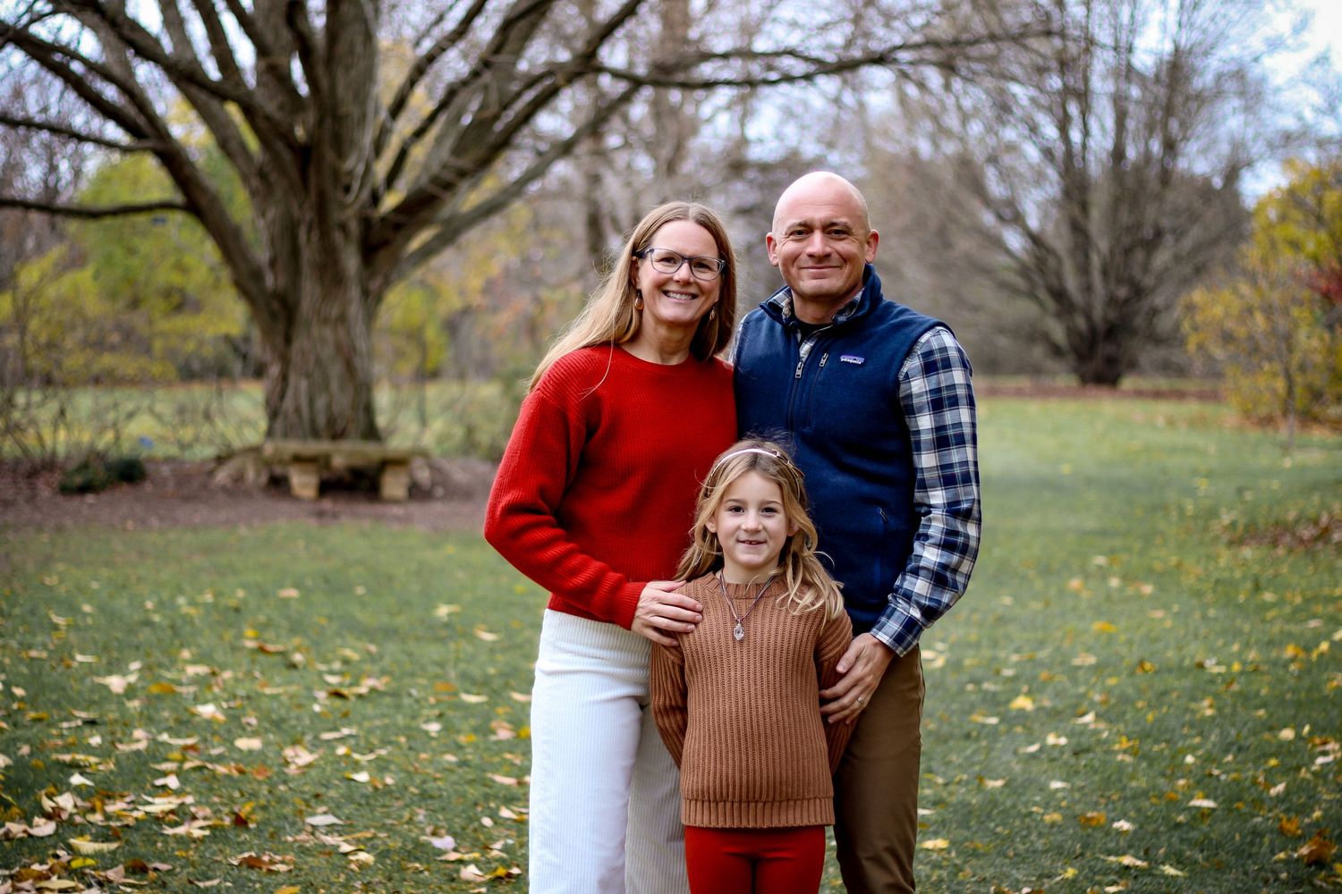 Family photo of a mom, dad, and daughter in an open field with a large tree behind them