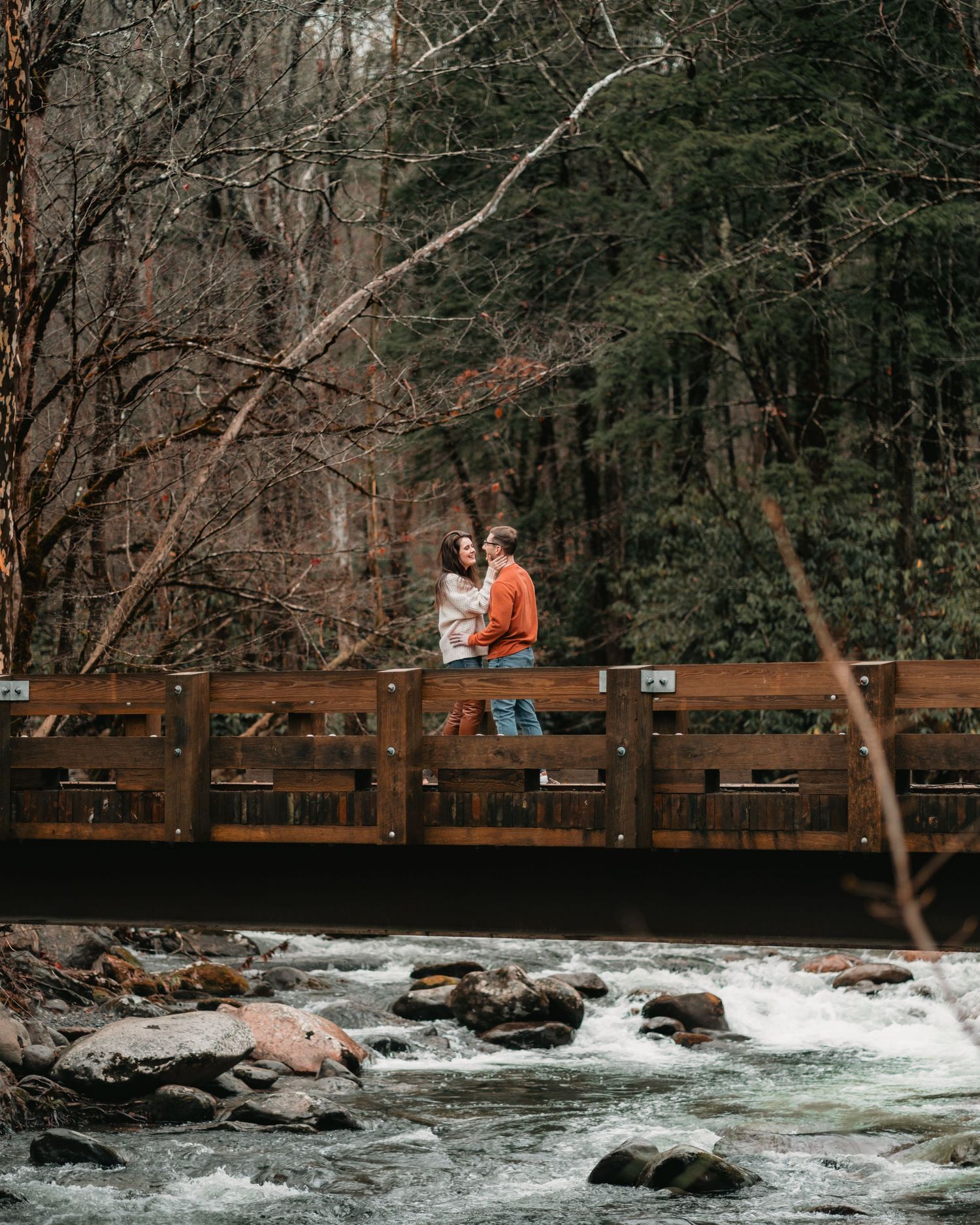 A couple sharing an intimate moment on a wooden bridge over rushing water in a forest setting.