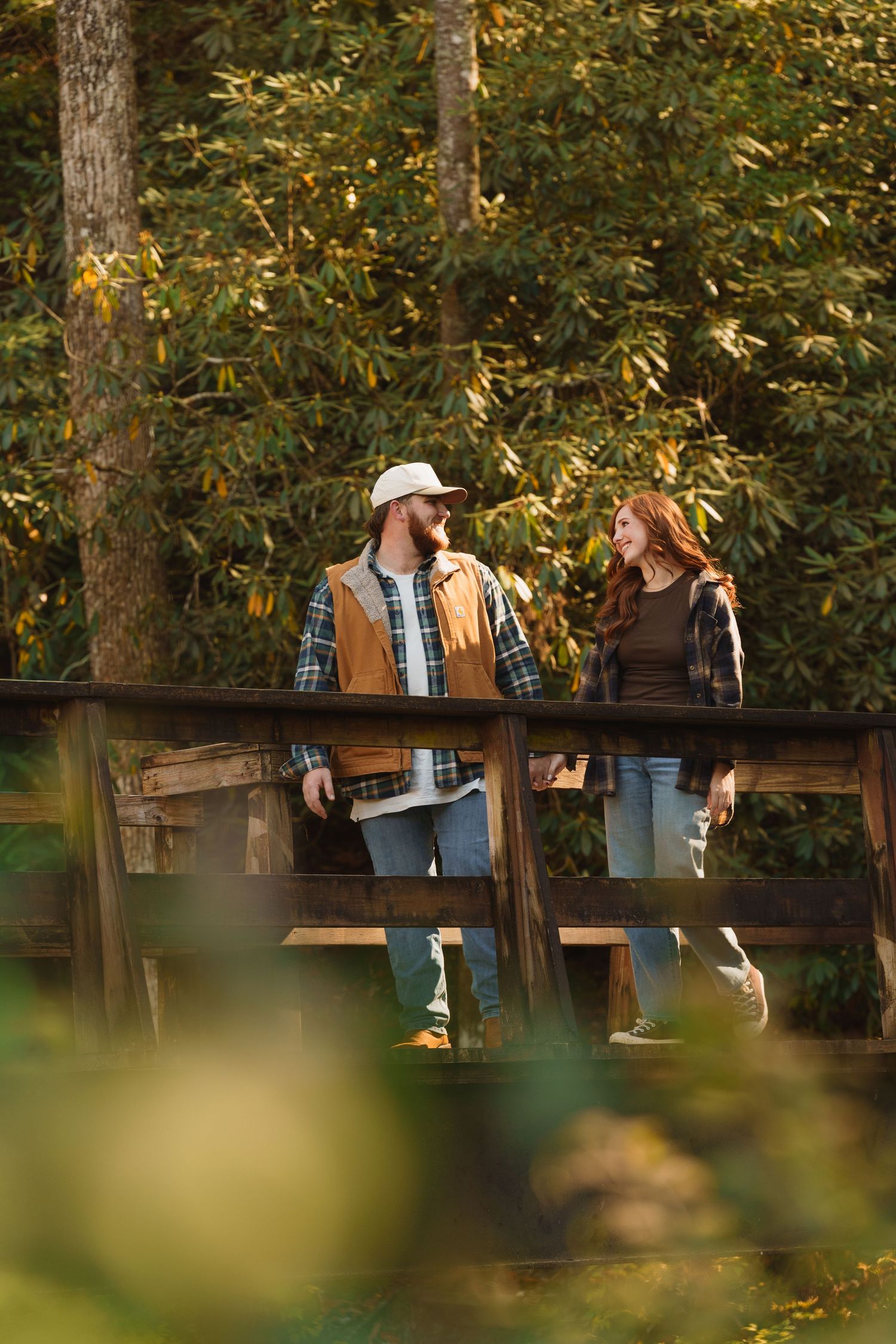 Couple standing on a wooden bridge surrounded by lush green forest trees during golden hour.