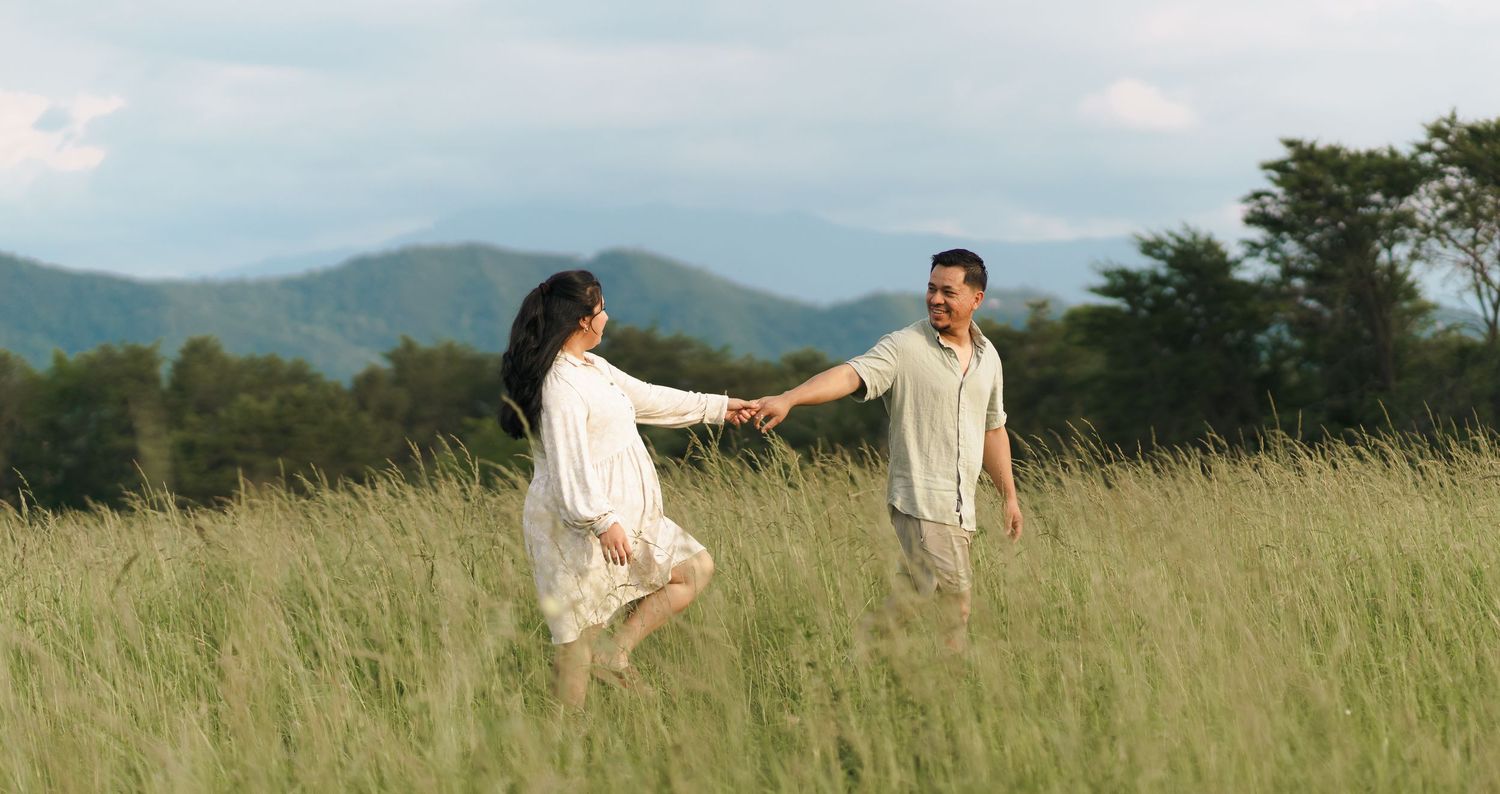 A couple in white outfits dances together in a scenic mountain meadow with tall grass and cloudy skies.