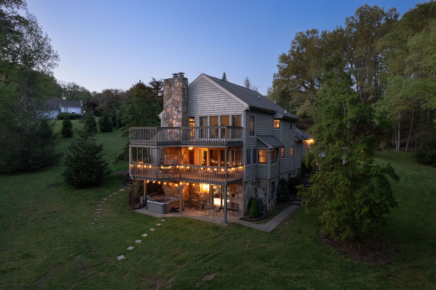 Modern three-story lake house with wraparound deck and string lights glows warmly at dusk among trees.