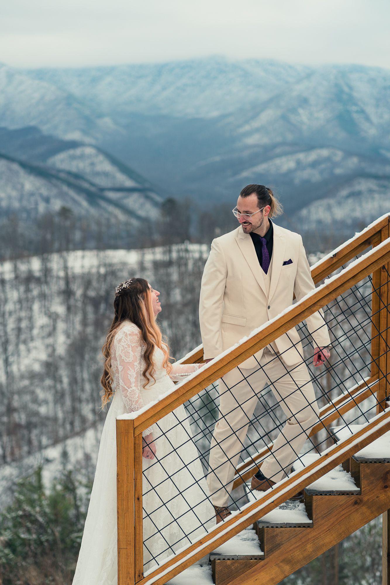 A couple in winter wedding attire stand on wooden stairs overlooking snowy mountain peaks in a romantic winter setting.