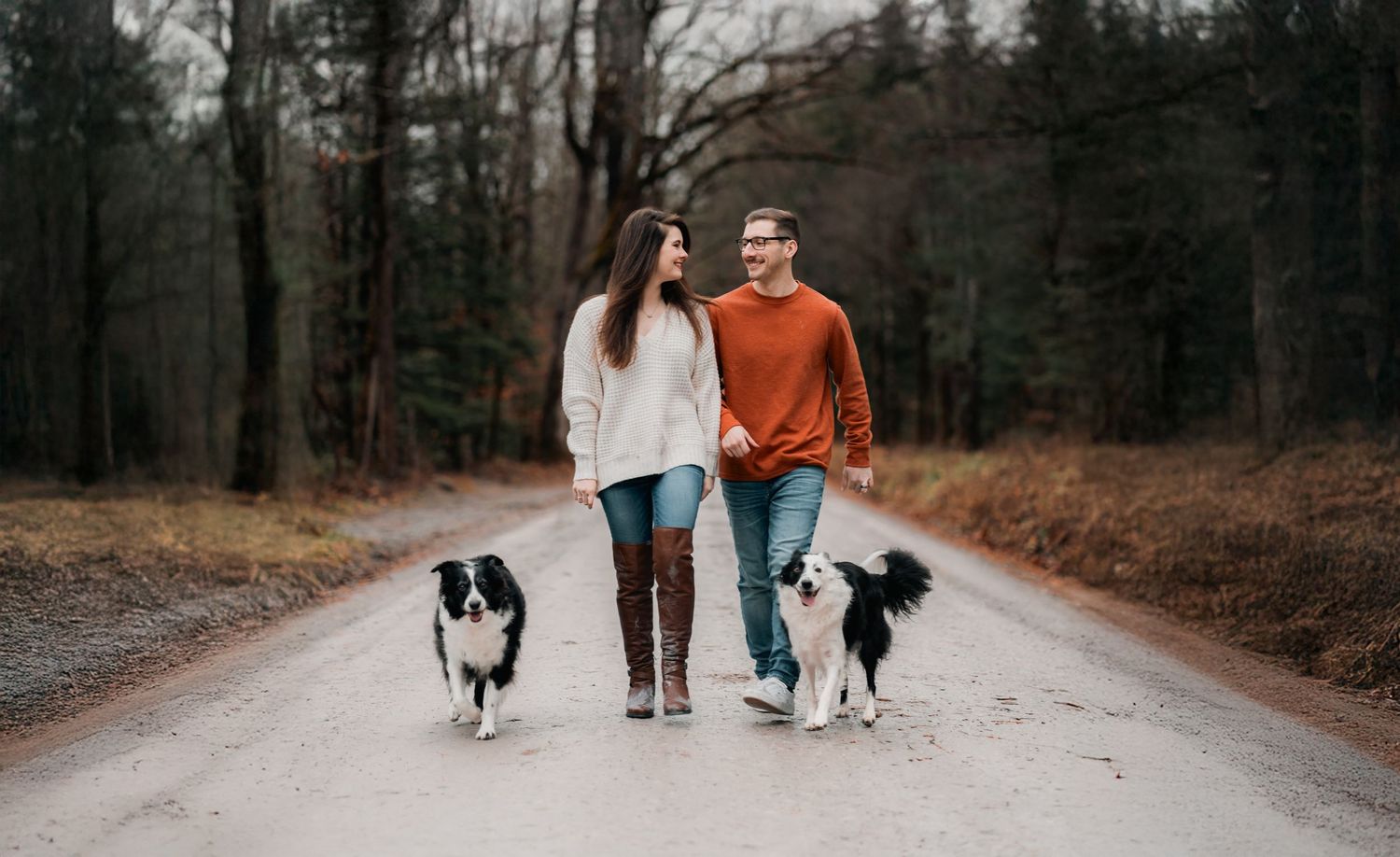 A couple walks their two border collies down a wooded path on an autumn day.
