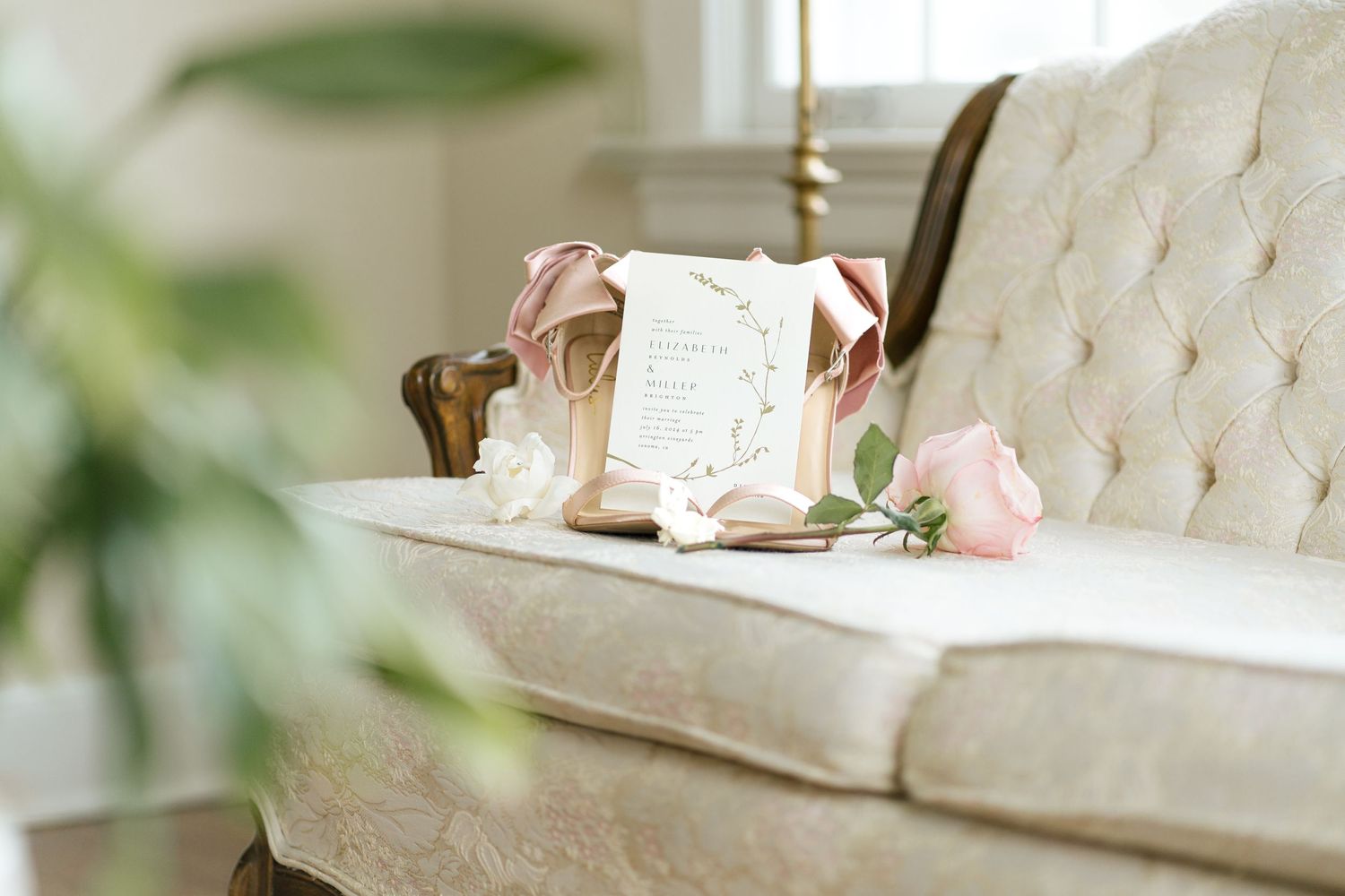 An elegant wedding invitation displayed on a cream tufted sofa with soft pink roses.
