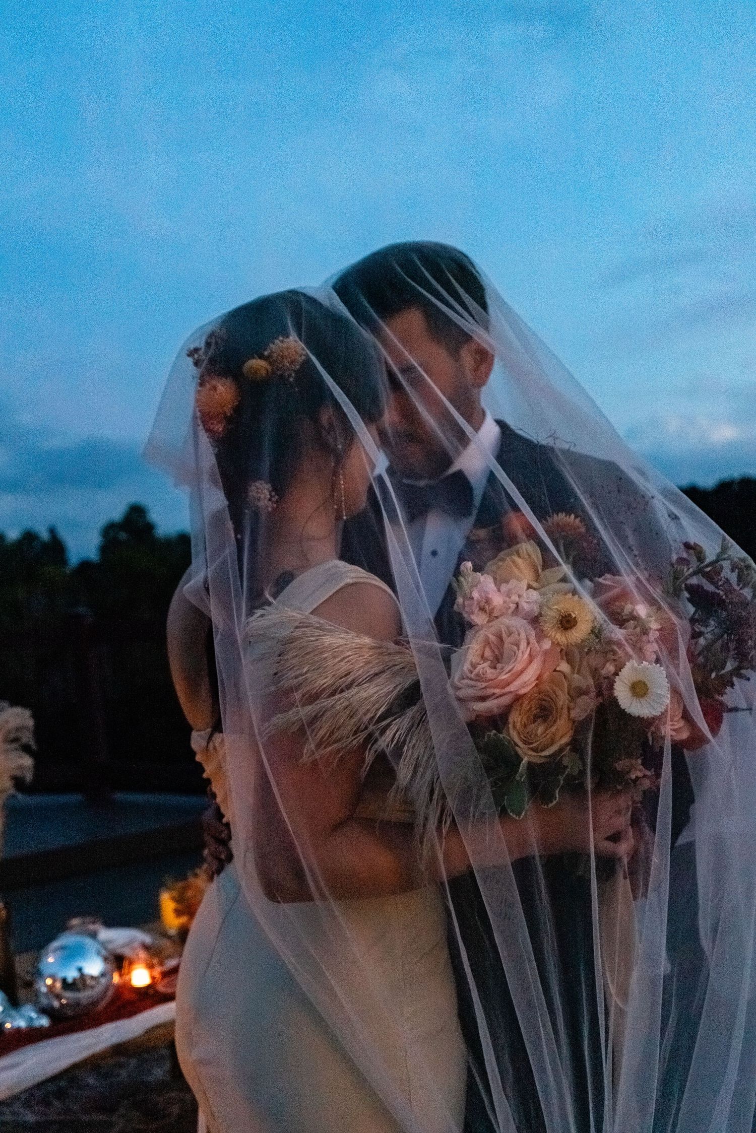 Romantic sunset wedding portrait under flowing veil with pink floral bouquet.
