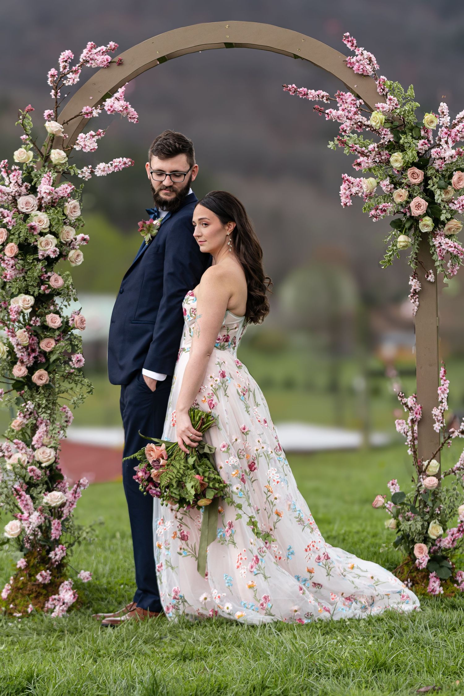 Couple poses under pink floral wedding arch in elegant outdoor ceremony setting.
