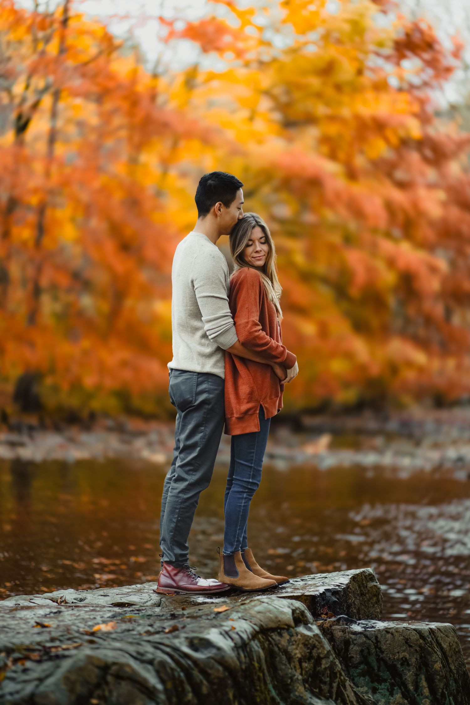 A couple stands together on a fallen log over a stream surrounded by vibrant autumn foliage.