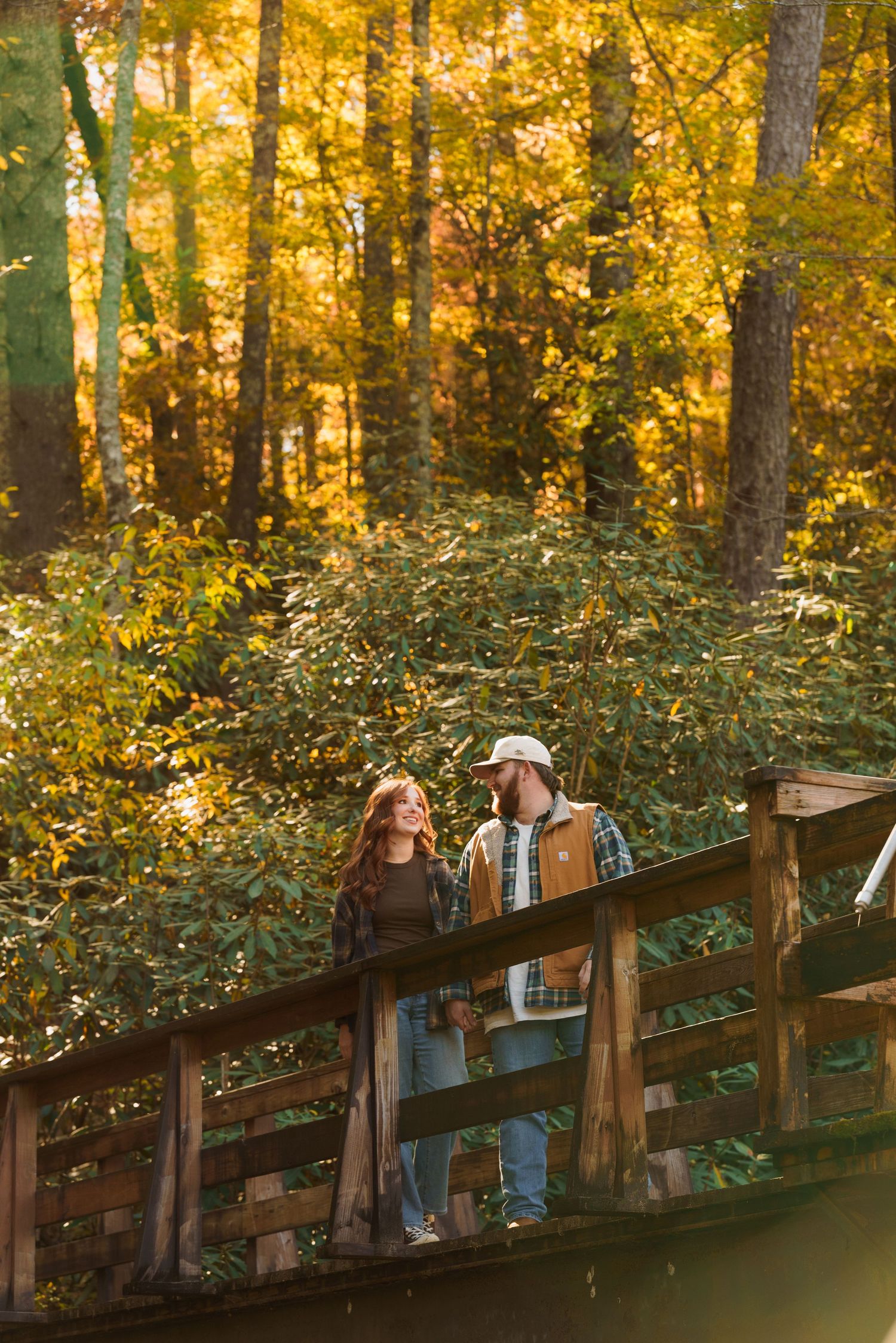 Two people stand on a wooden footbridge surrounded by vibrant golden autumn foliage in a sunlit forest.