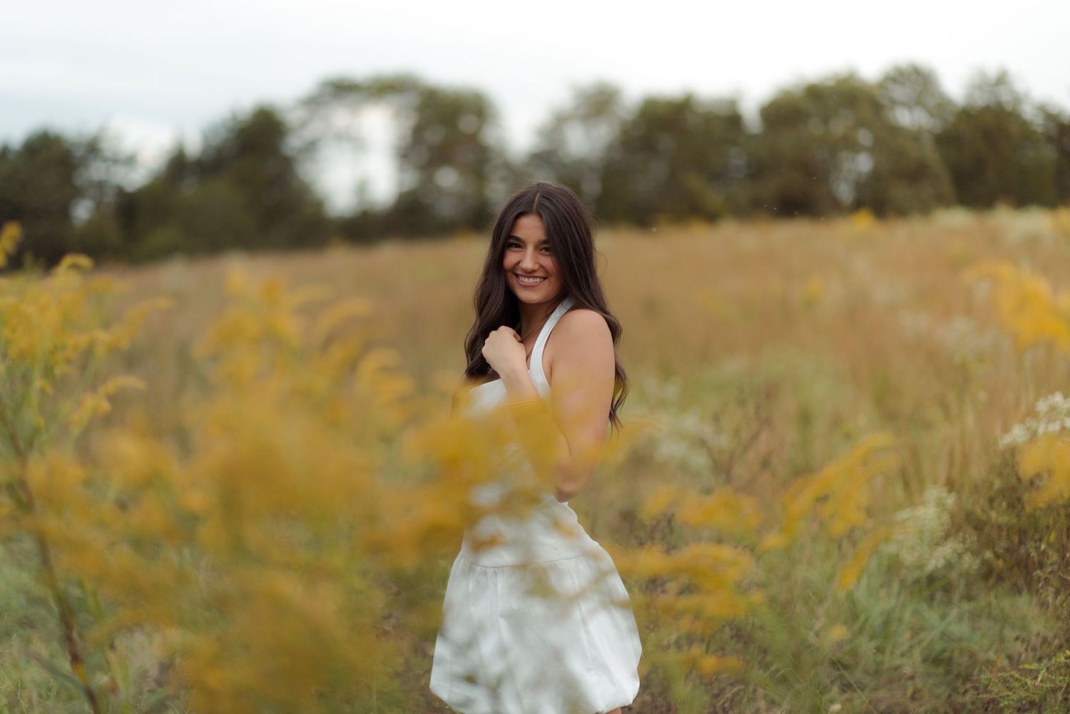 A figure in a white dress stands in a golden field of wildflowers during sunset, turning to look over their shoulder.
