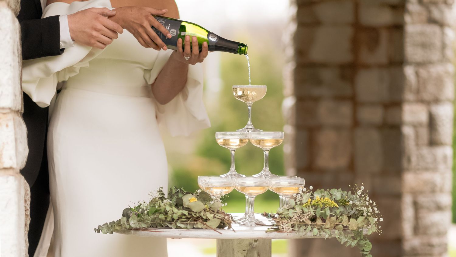 Champagne being poured into a tower of glasses decorated with greenery.