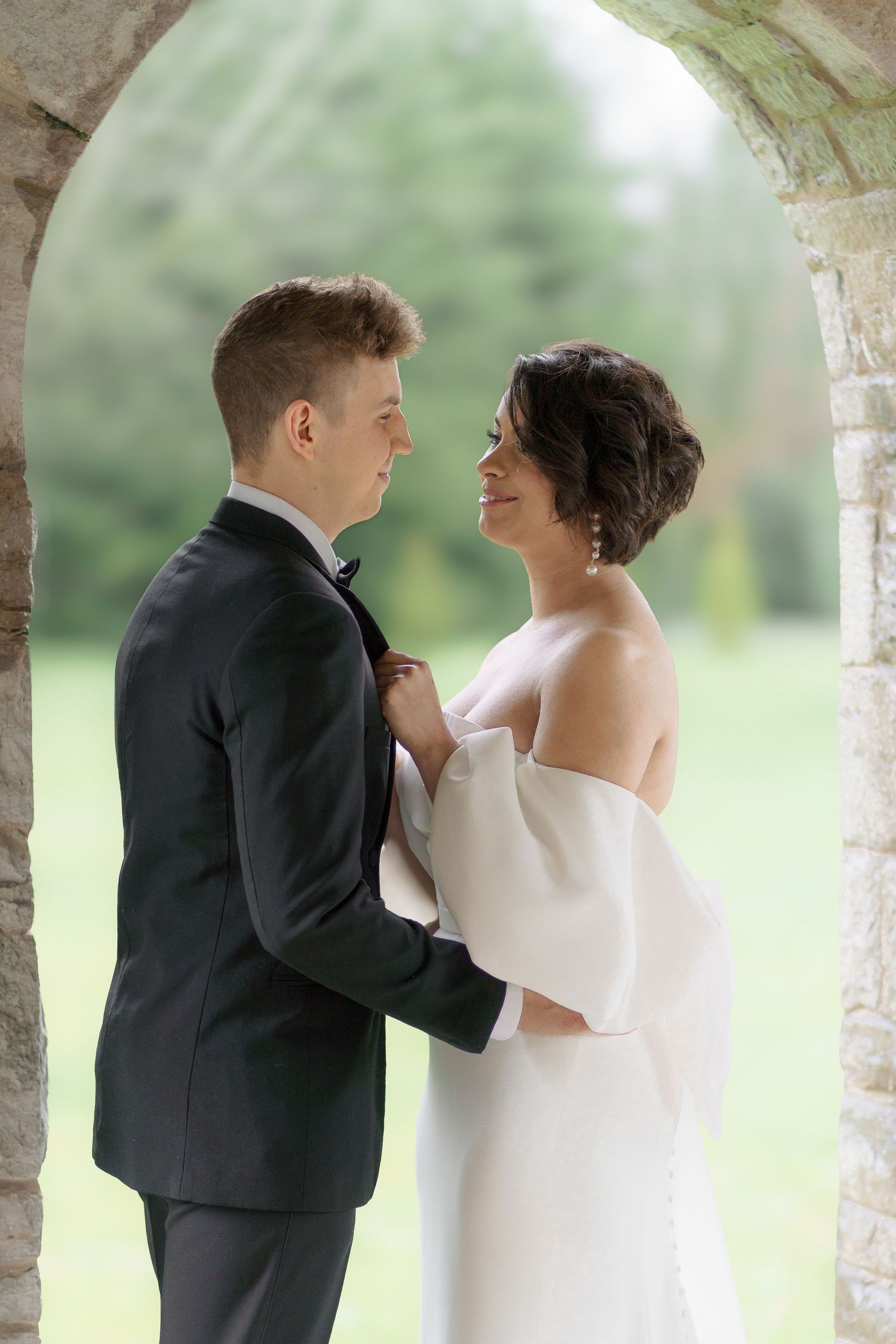 A couple shares an intimate moment under a stone archway in formal wedding attire.
