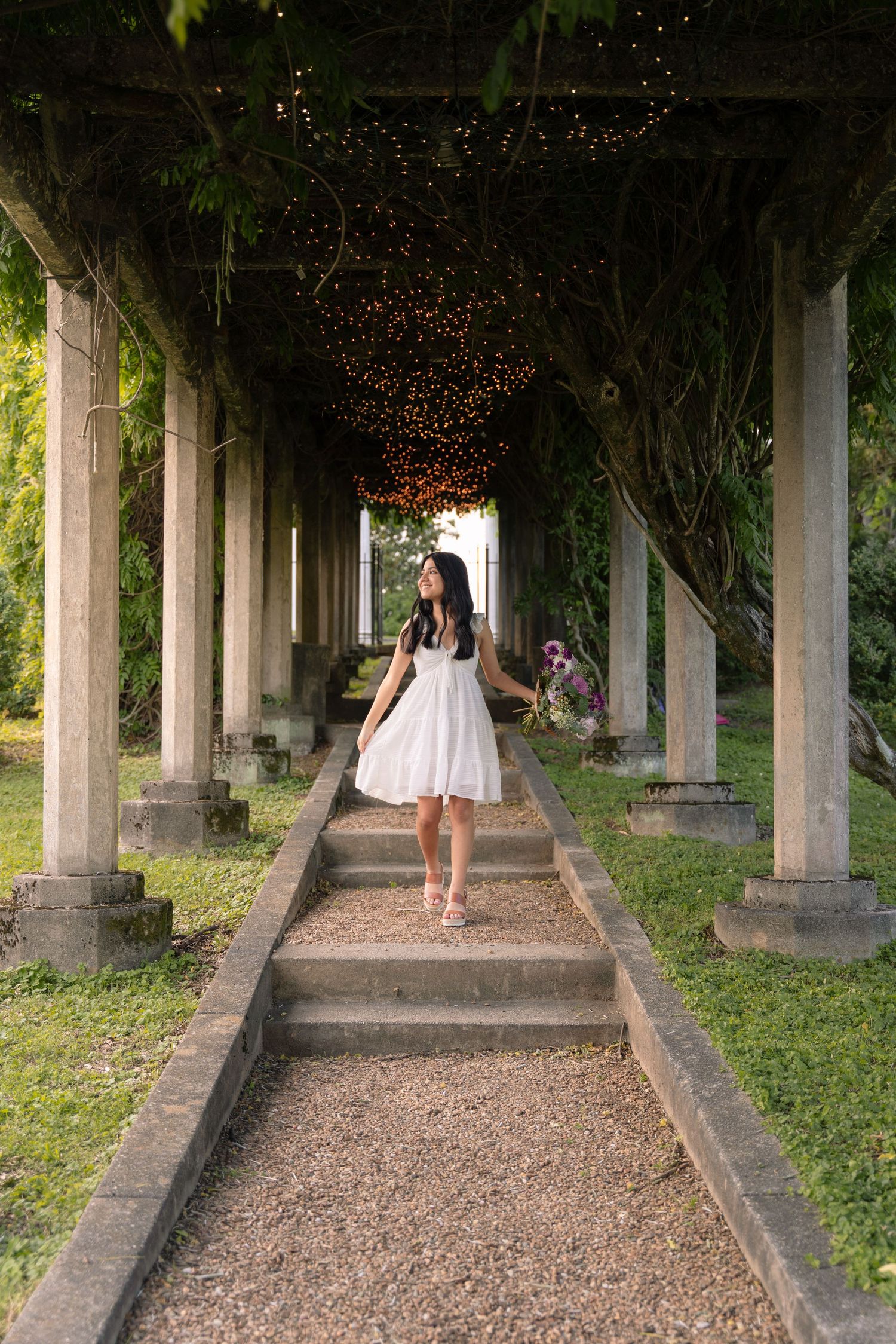 A person in a white dress stands on garden steps under string lights and columns.