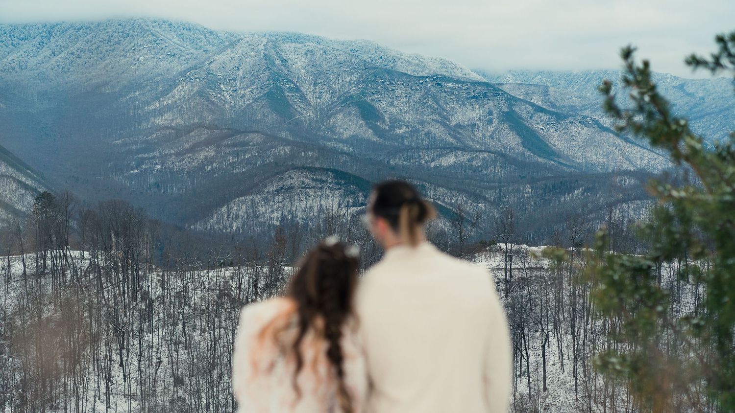 A couple admires the snowy mountain vista from a scenic overlook in winter.