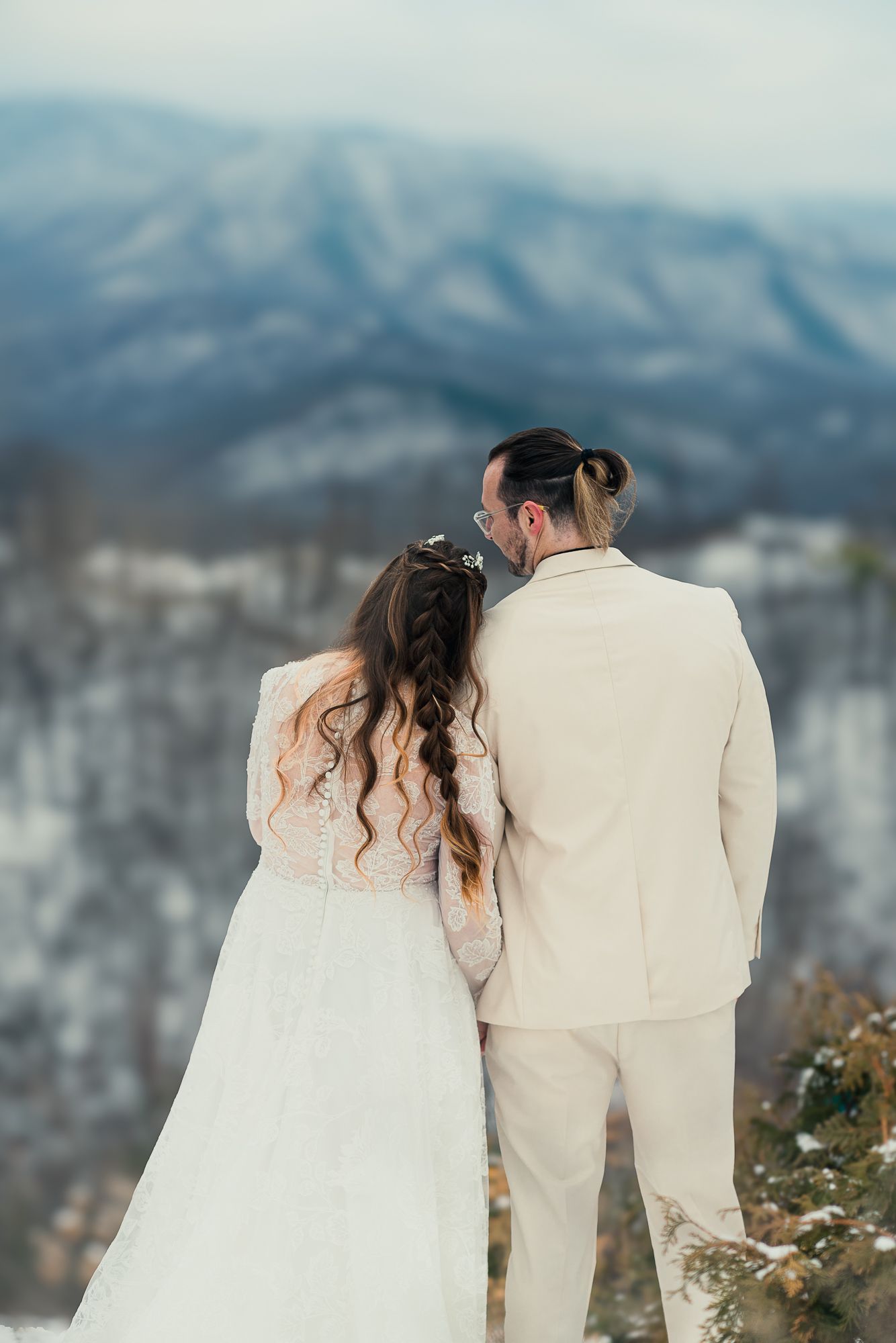 A couple in white wedding attire stand together overlooking snowy mountain peaks on their special day.