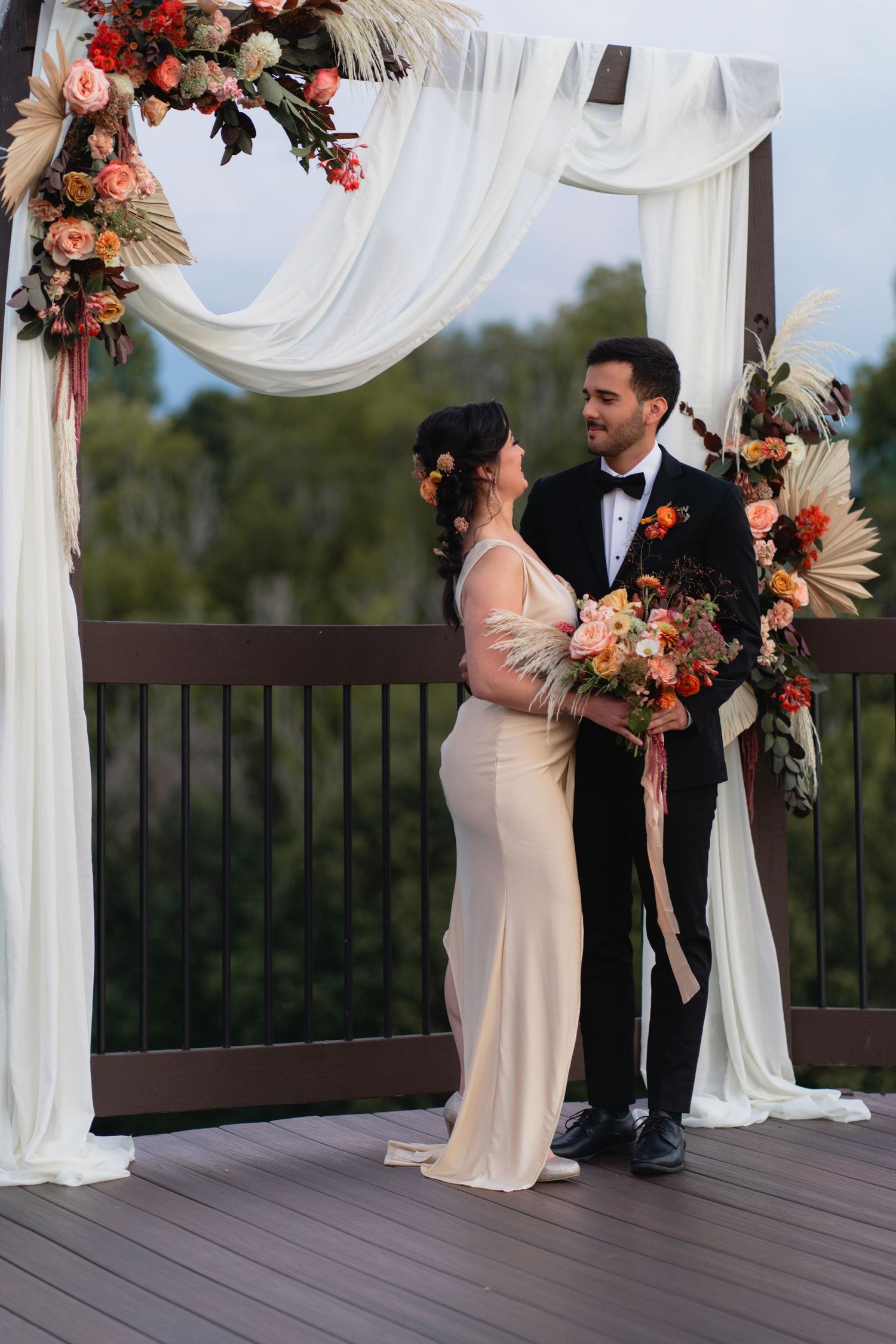 A wedding ceremony under a draped arch decorated with coral and peach flowers on an outdoor deck.