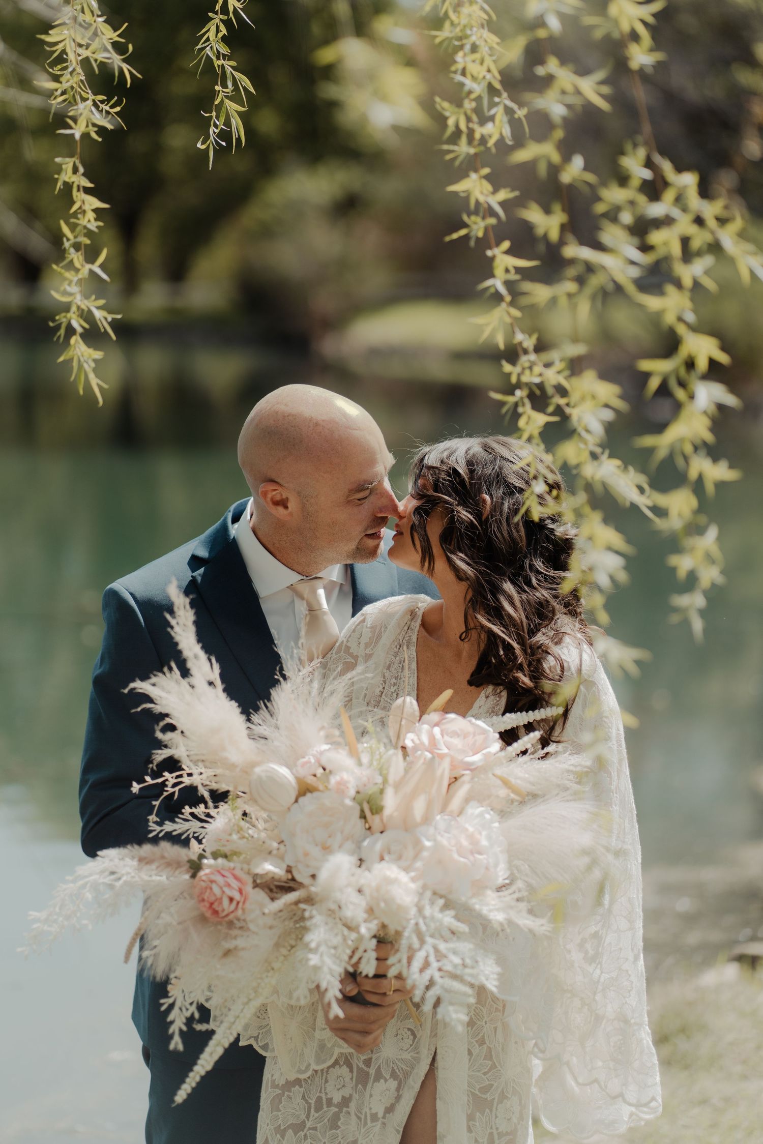 Couple sharing intimate wedding moment with pampas grass bouquet by water.