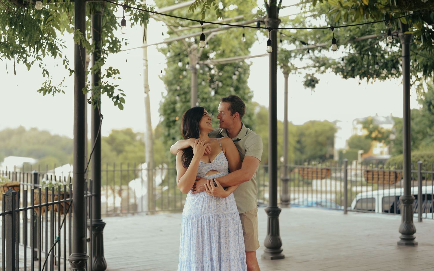 A couple embraces romantically under a vine-covered pergola on a scenic waterfront promenade at sunset.