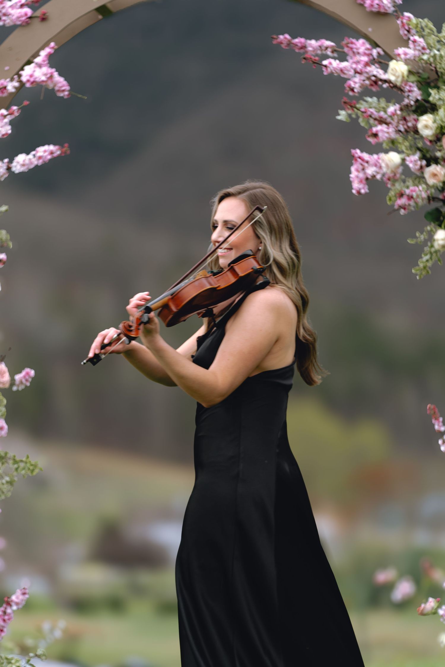 Violinist in black dress performs under cherry blossoms with mountains in background.