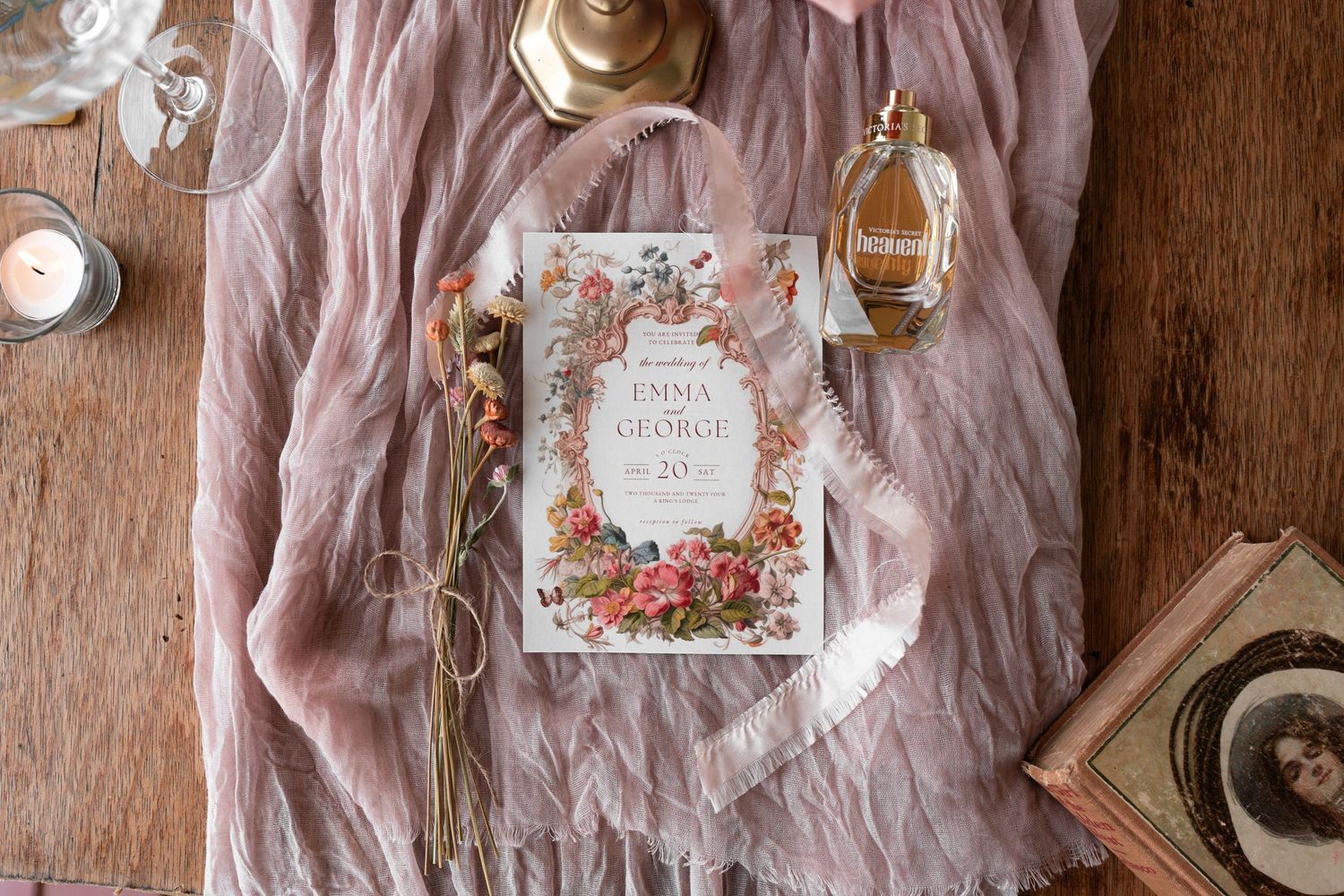 Wedding details flatlay with pink fabric, perfume bottle and floral invitation book.