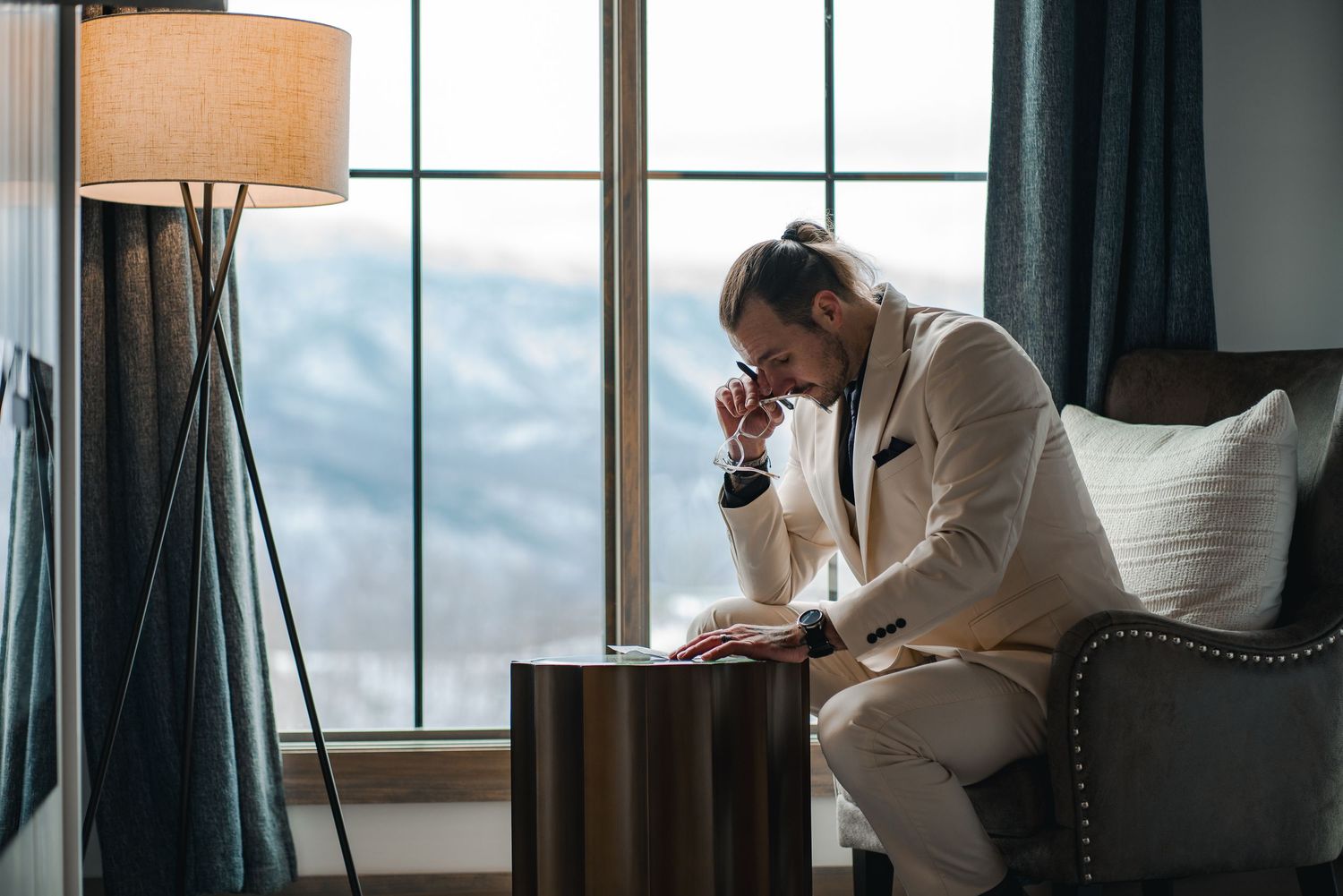 A person in a white suit sits contemplatively near a window in a hotel room with a modern floor lamp.