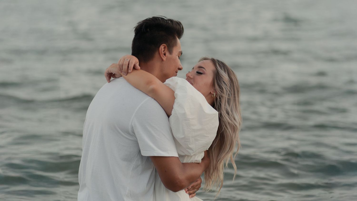Romantic embrace by the ocean during sunset with soft lighting.
