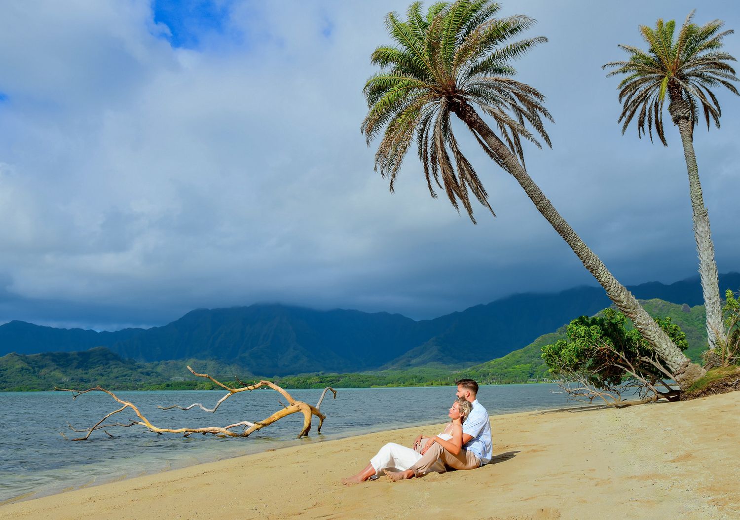 Couple sitting on a tropical beach with palm trees, mountains, and calm water in the background.