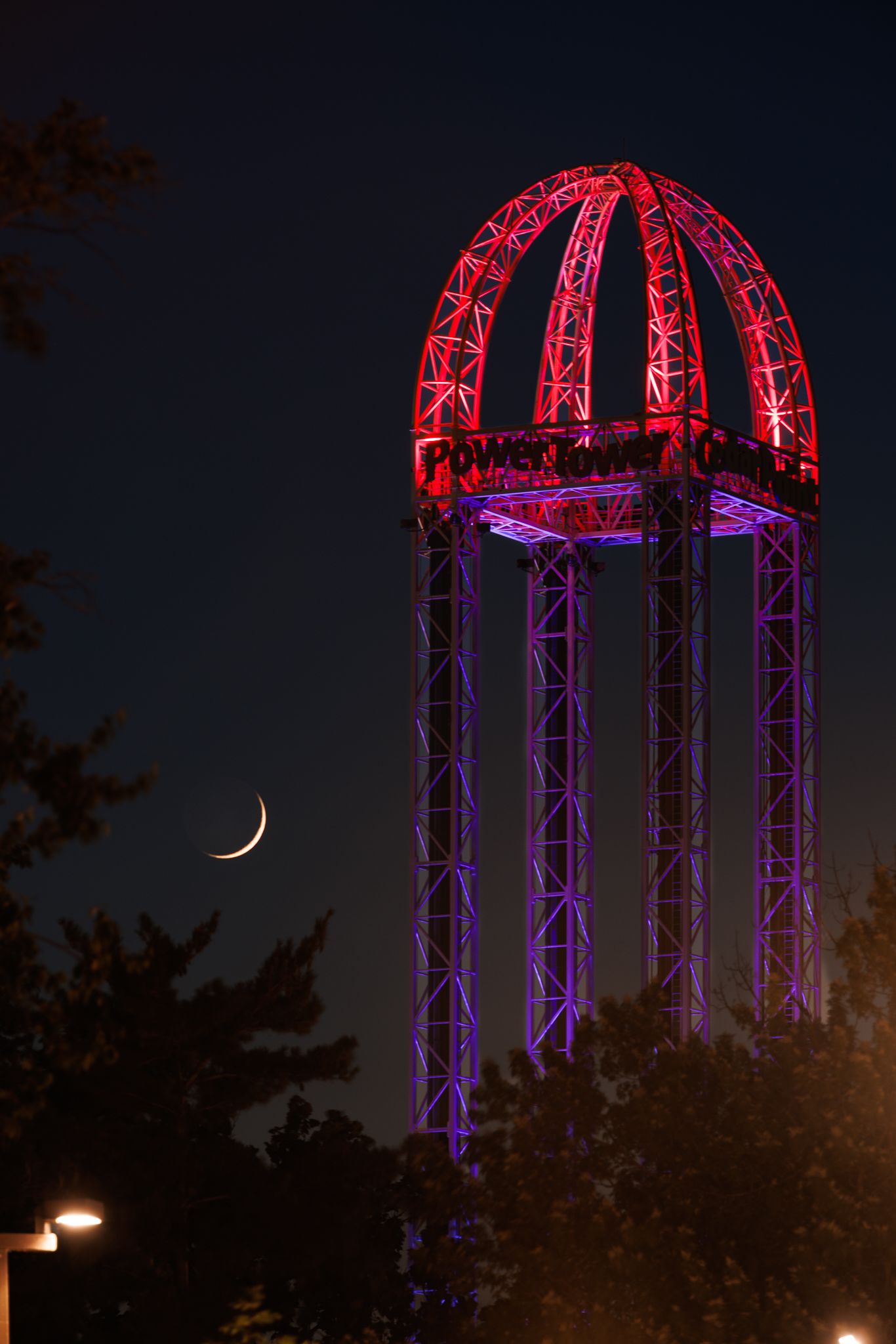 Illuminated red and purple lattice water tower at night with a crescent moon visible in the dark blue sky.