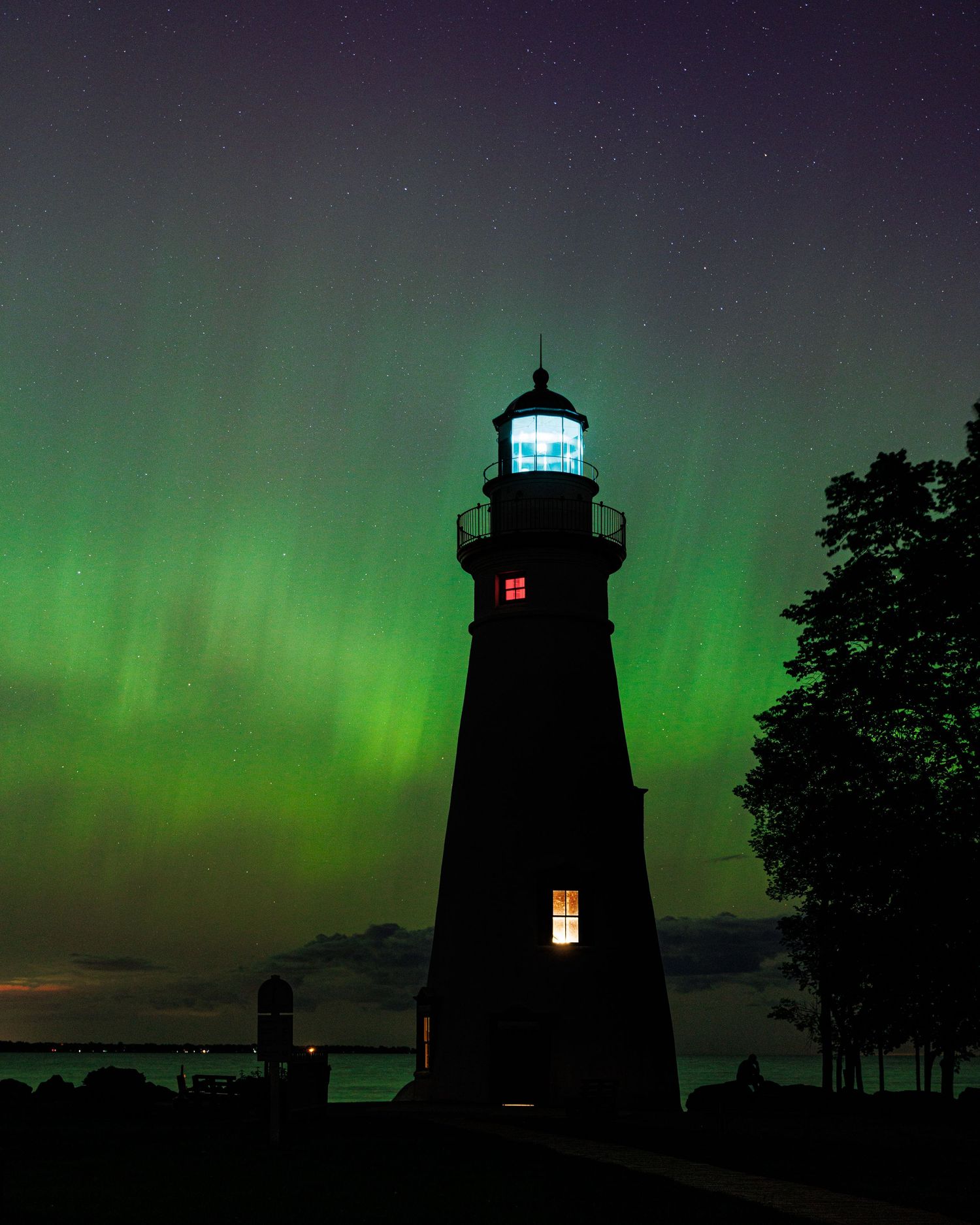 Lighthouse silhouette glowing against vibrant green northern lights aurora borealis in a starry night sky.