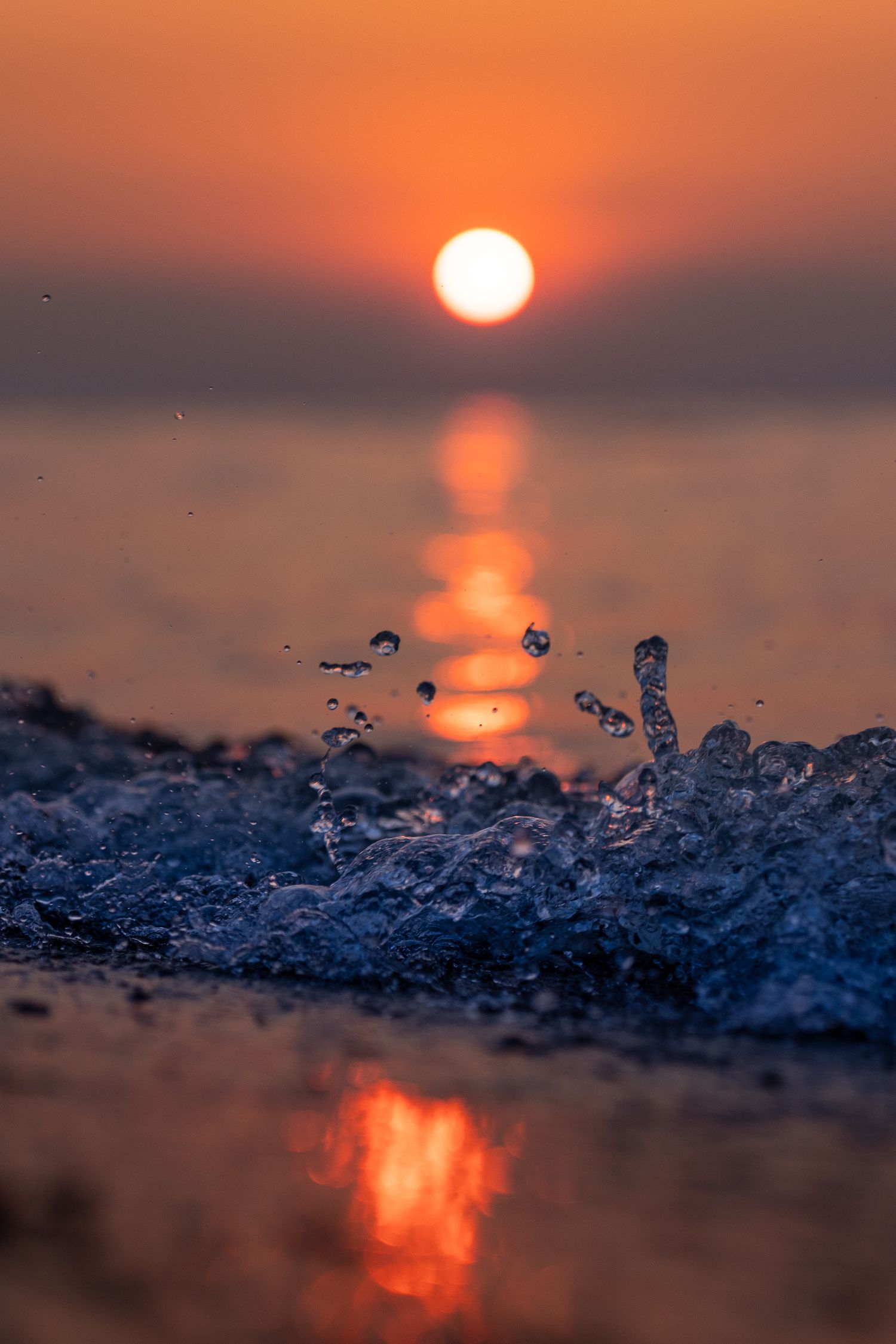 Close-up of ocean waves splashing at sunset with vibrant orange sky and sun reflecting on calm water.