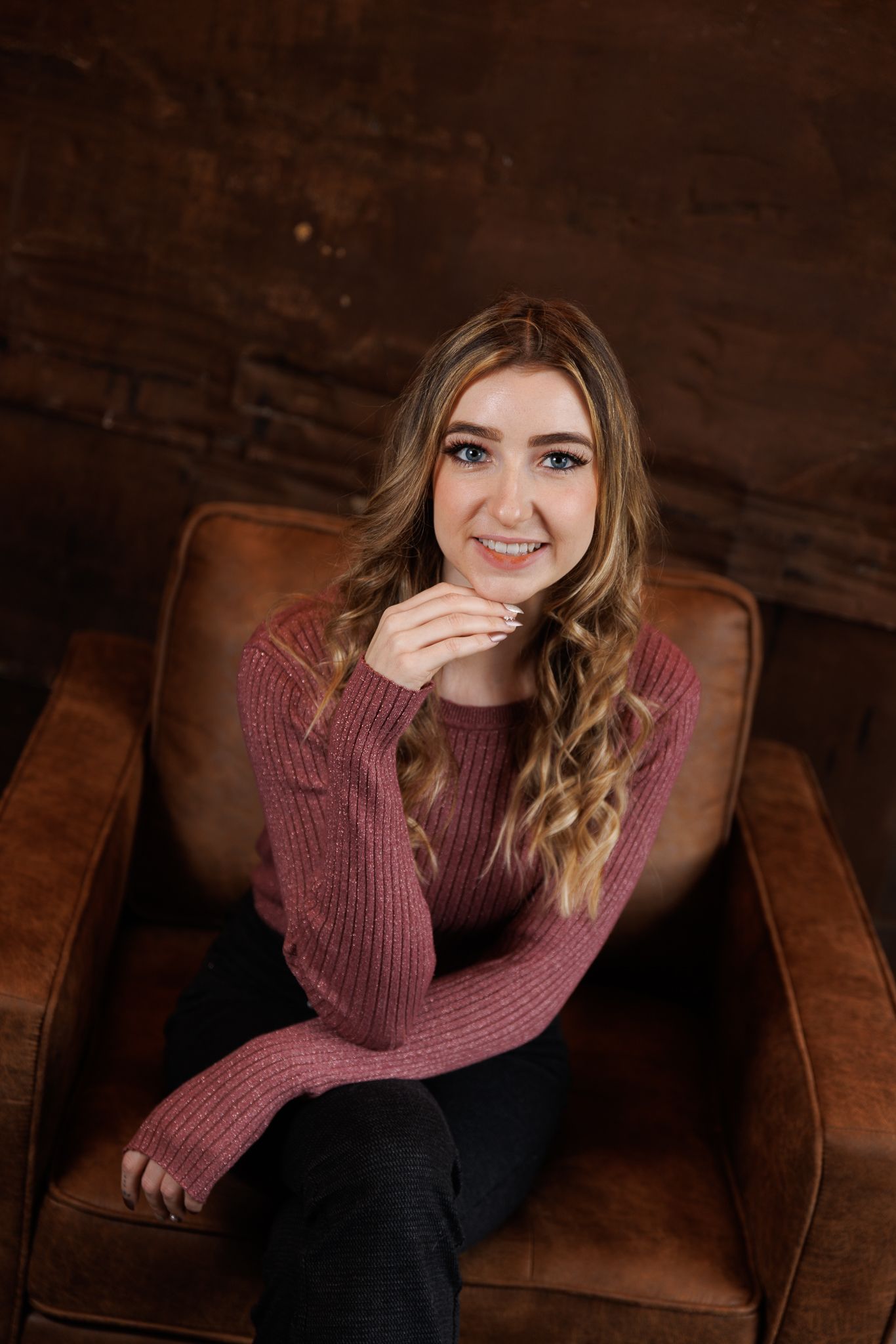 Young woman with wavy blonde hair smiles in a brown leather chair, wearing a pink knit sweater and black jeans.