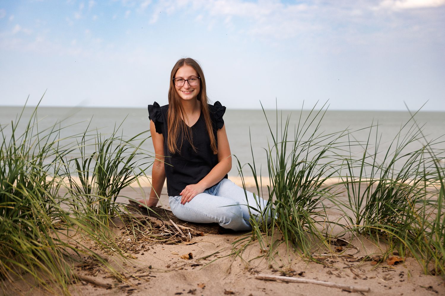 A person sits casually on a sandy beach dune with tall grass and ocean views in the background.
