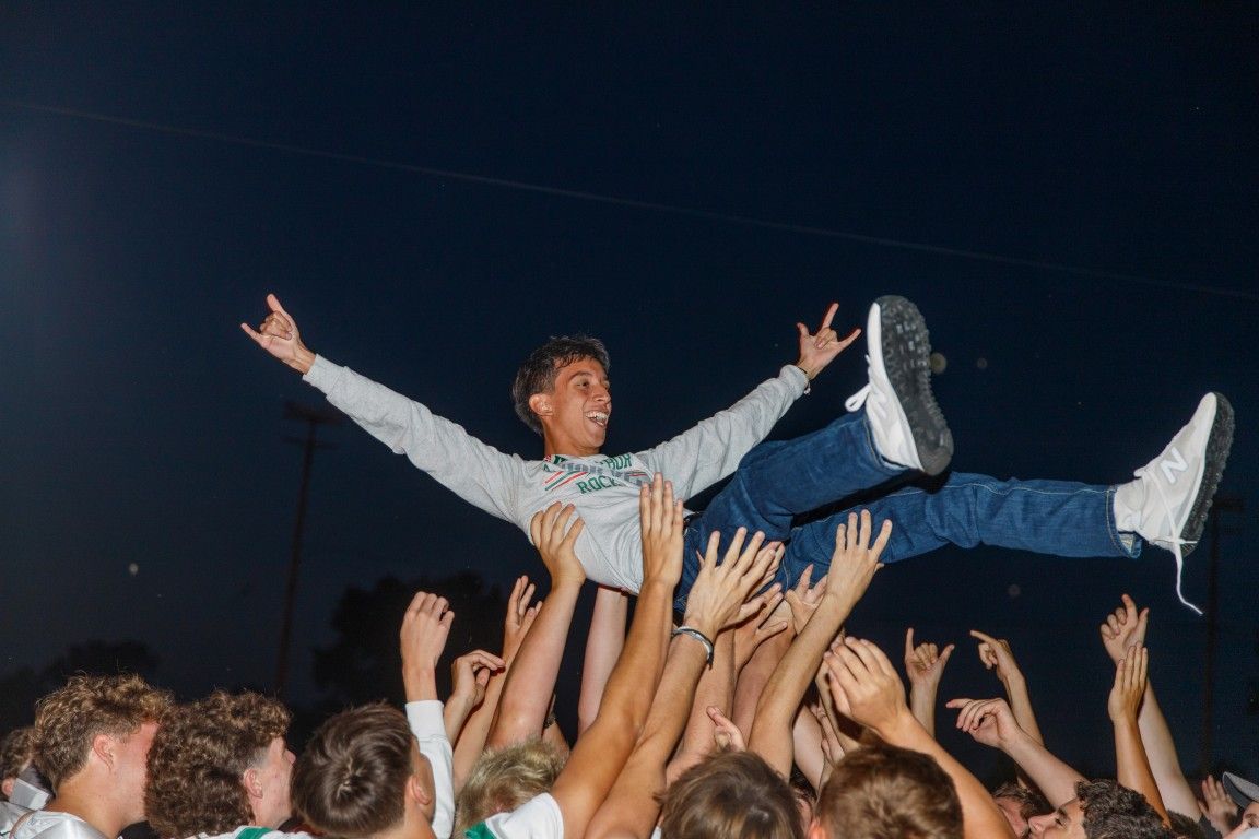 Young man crowd surfing at an outdoor concert, arms outstretched, wearing white hoodie and blue jeans.