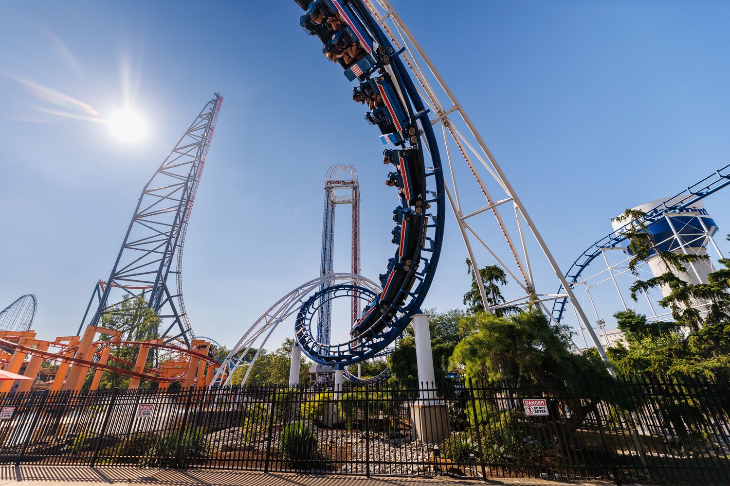Thrilling roller coaster loop at an amusement park under bright blue sky with lush green trees surrounding the ride.