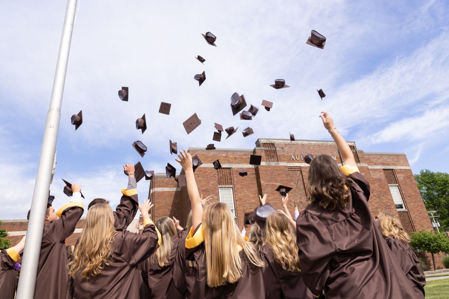 Graduates in brown caps and gowns tossing mortarboards into blue sky during outdoor graduation ceremony.