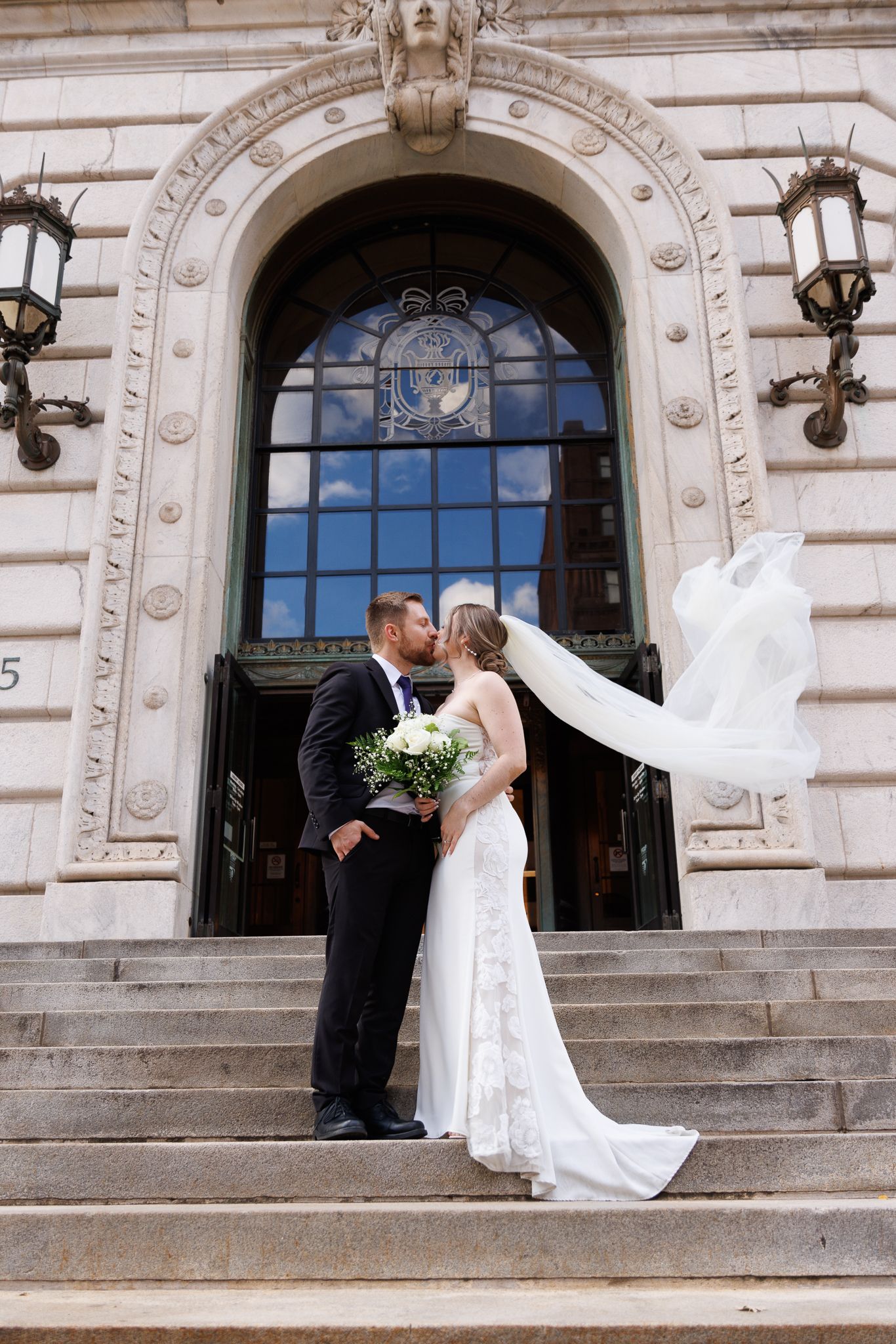 Bride and groom kissing on steps of elegant building, her veil flowing in wind, holding white bouquet.