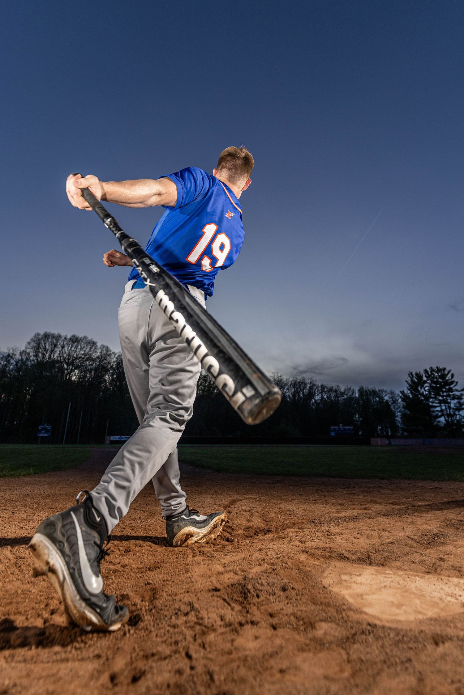 Baseball player #19 in blue jersey swings bat at home plate during evening game on dirt field.
